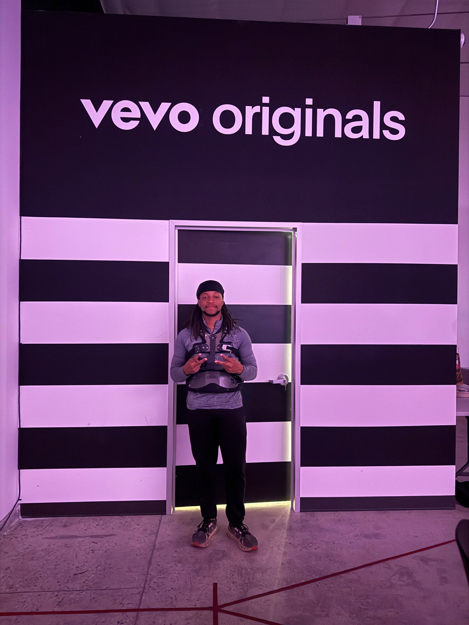 Person taking a mirror selfie in front of a black and white striped wall with 'vevo originals' sign, holding a phone, standing on a gray floor.
