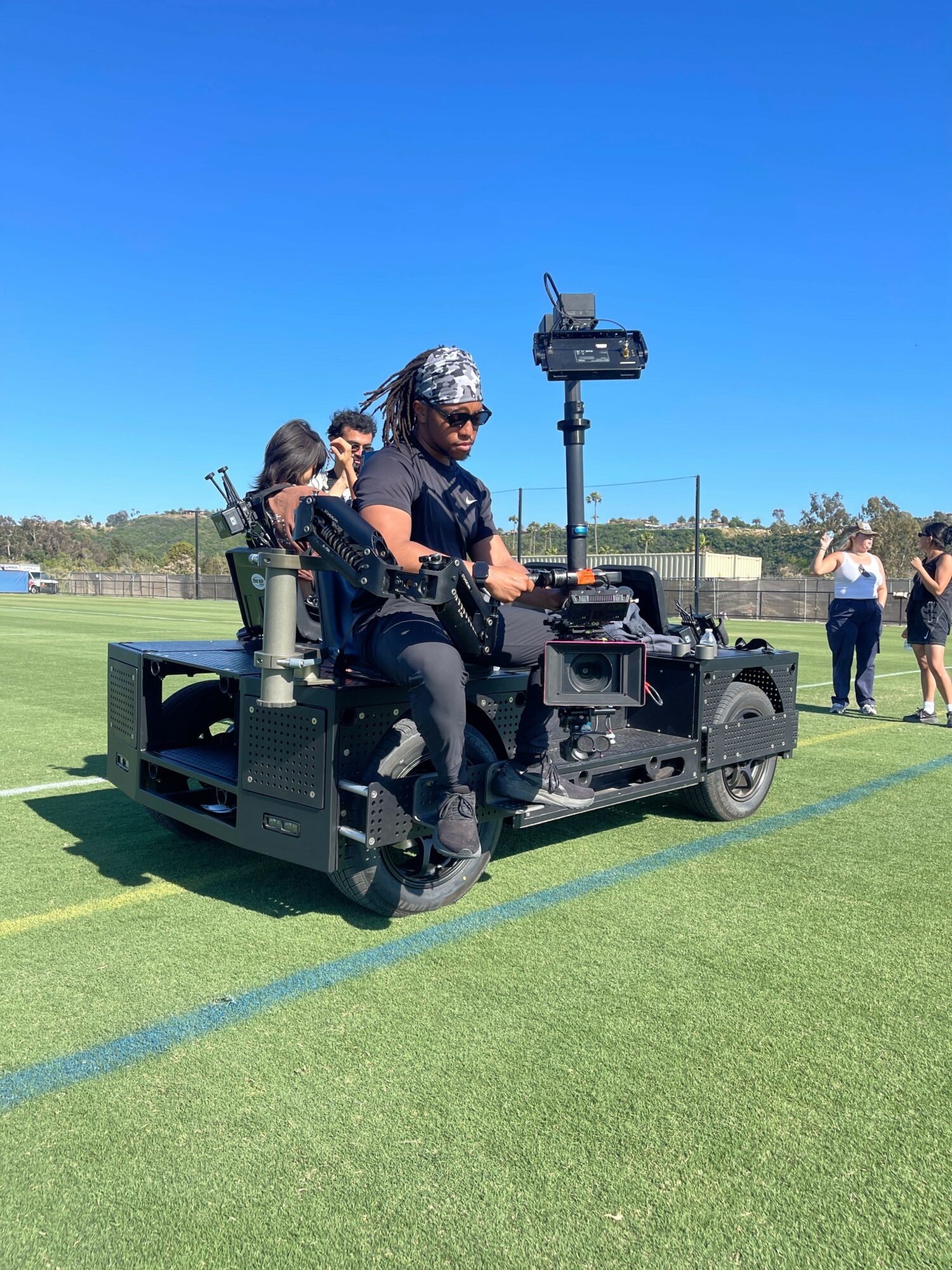 Person operating a large camera mounted on a vehicle on a grassy field with a clear blue sky.