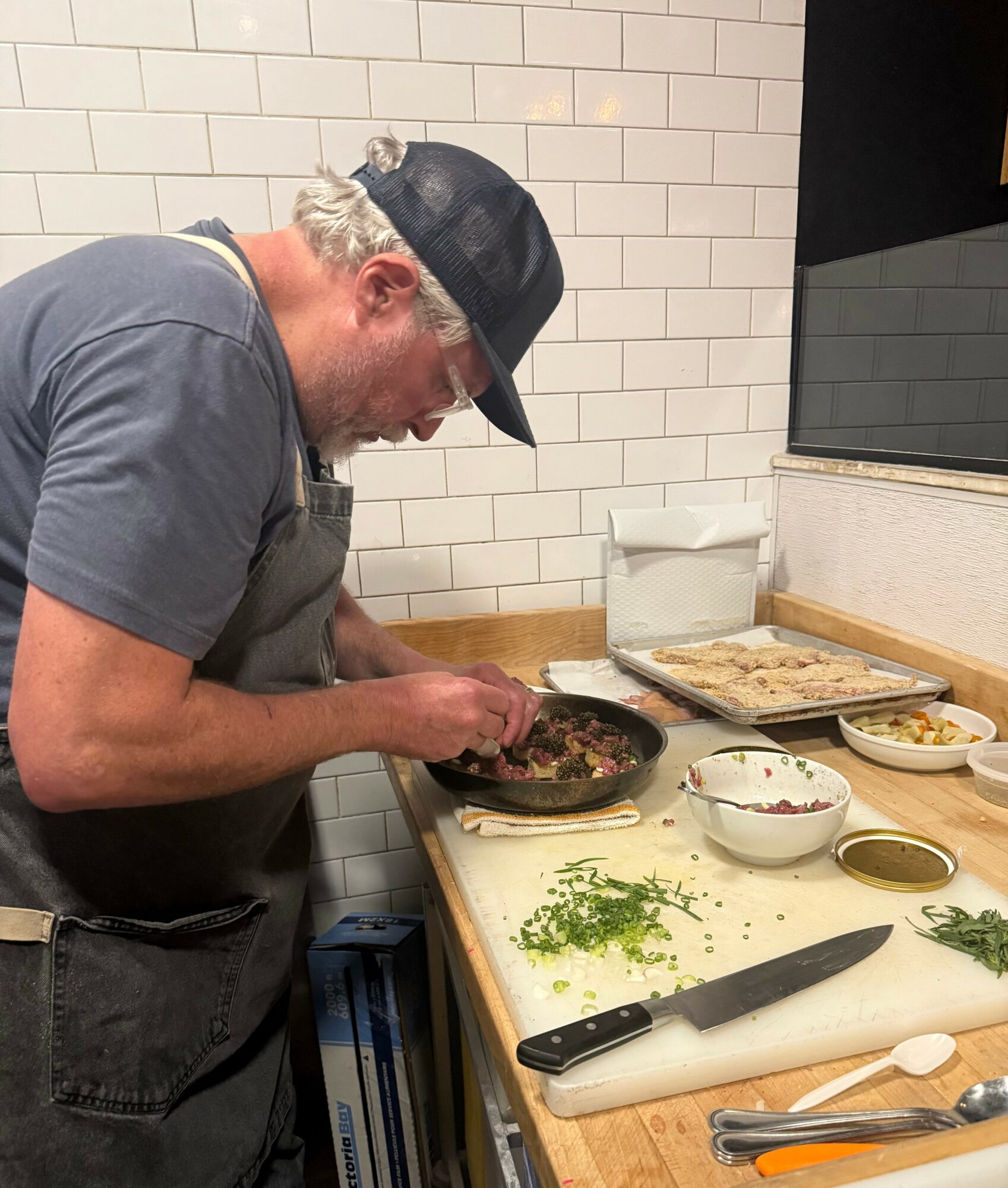 Man cooking in a kitchen, preparing food in a frying pan on a counter with bowls, a knife, and ingredients.