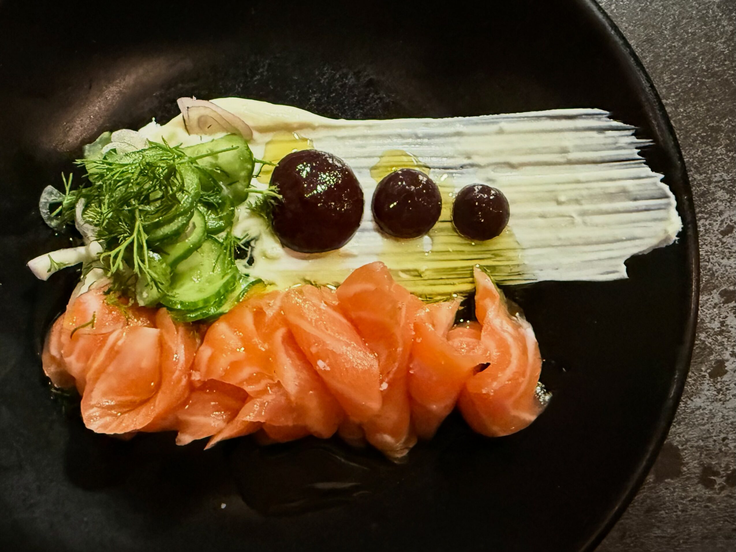 Slices of raw salmon, green vegetables, and mushrooms on a black plate.