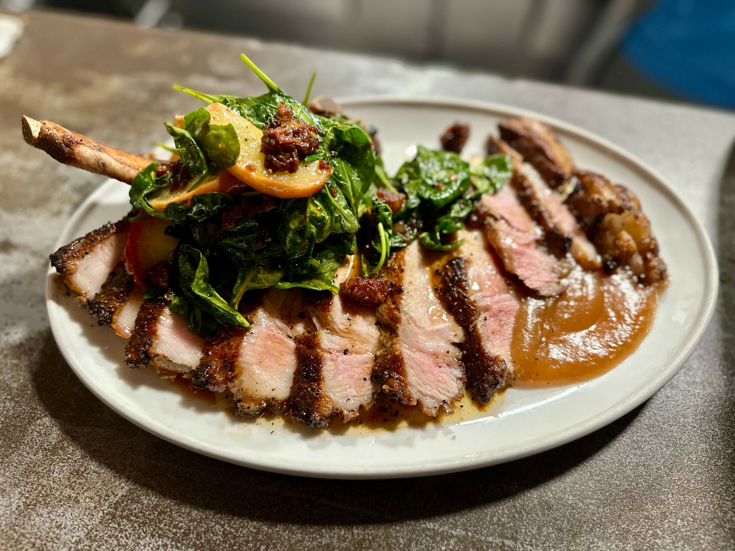 Sliced grilled steak with sauce, topped with greens and garnishes on a white plate.