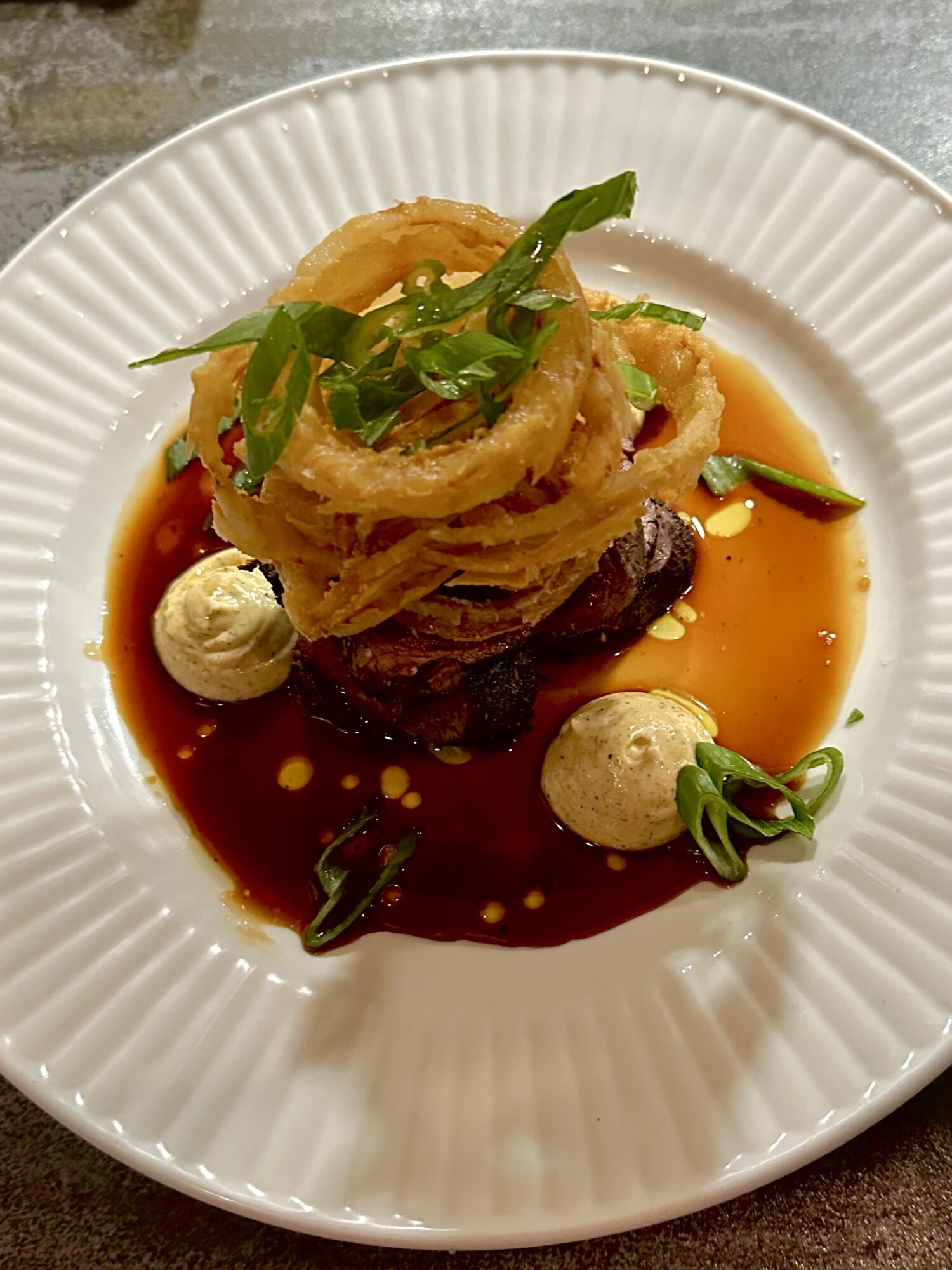 A plated dish with fried onion rings, green herbs, sauce, and small round garnishes on a white plate.