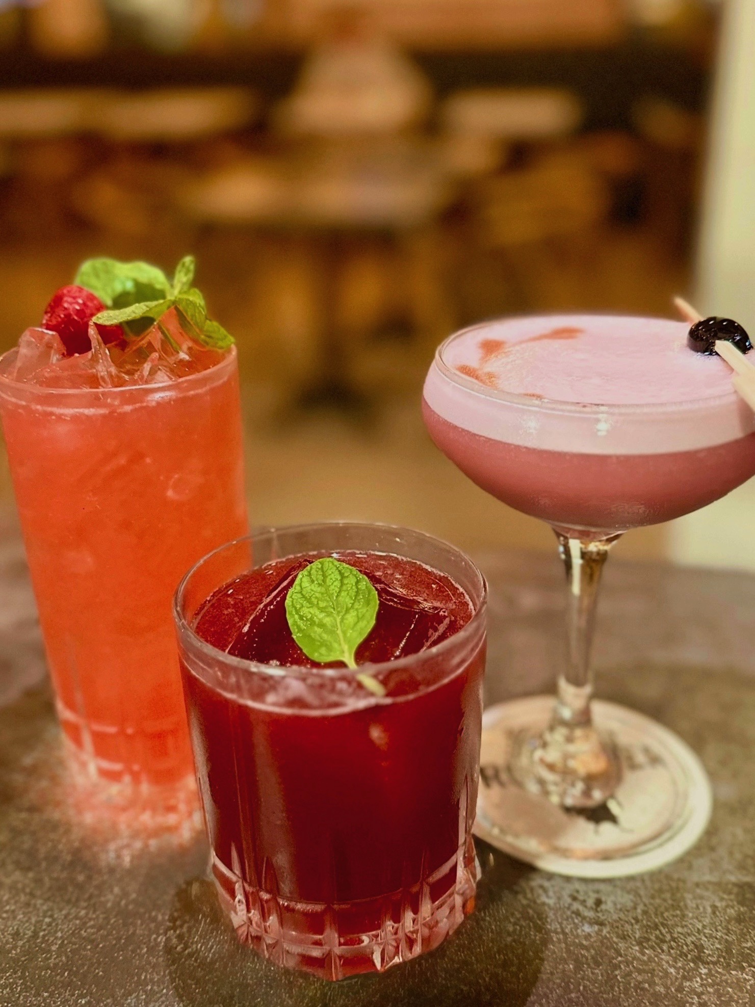Three colorful cocktails on a table, garnished with mint leaves and berries, with a blurred background.