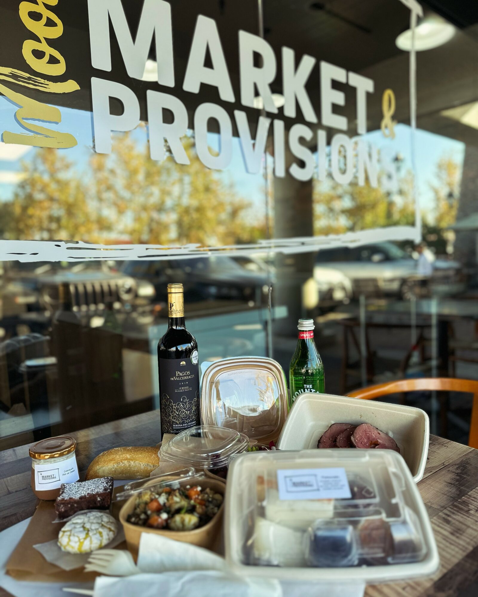 Display of food and drinks on a table inside a market, with a glass window reflecting trees and sky outside.