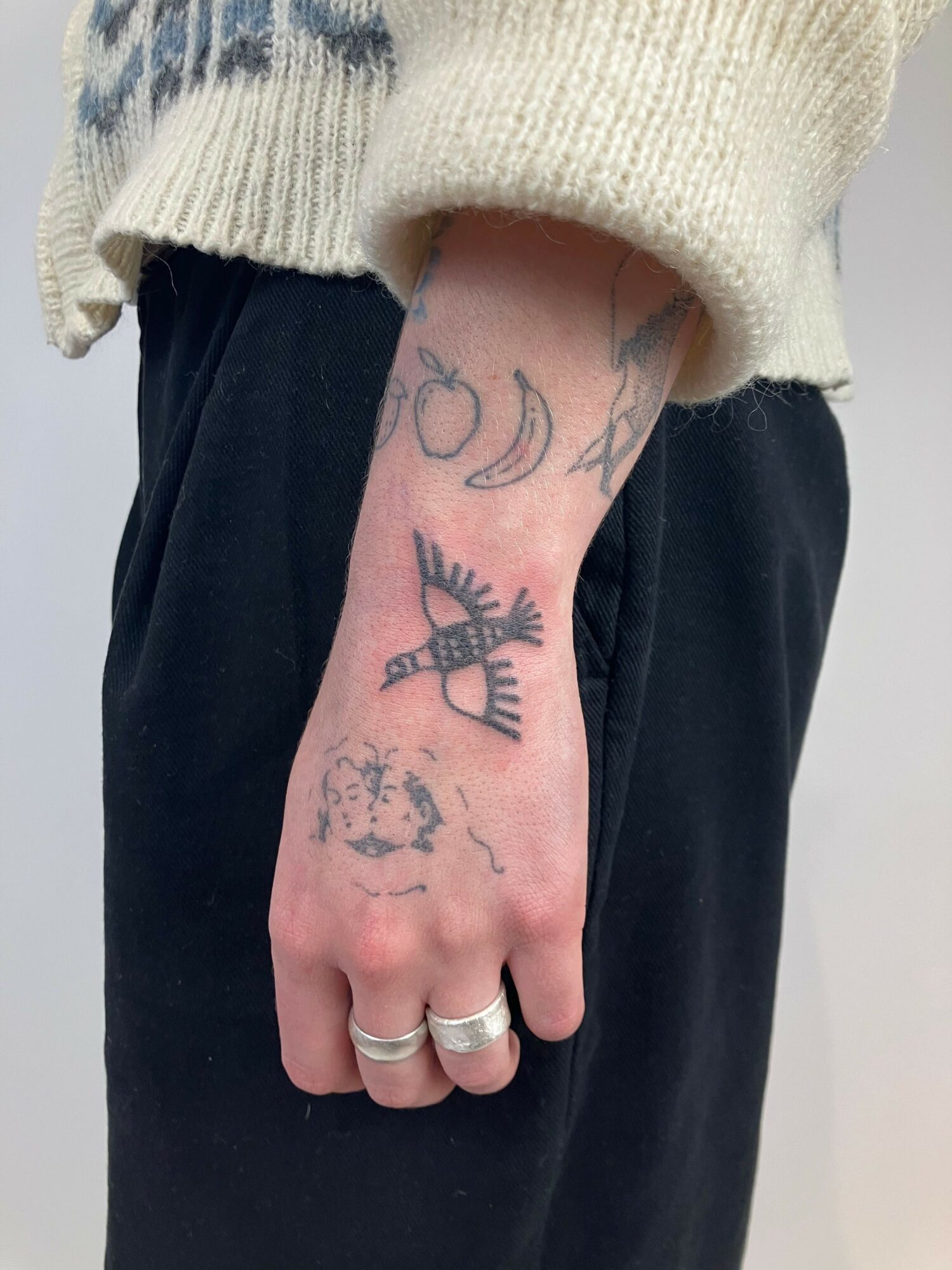 Hand with tattoos, wearing rings, resting on black pants, with a cream-colored sweater sleeve visible, against a plain background.