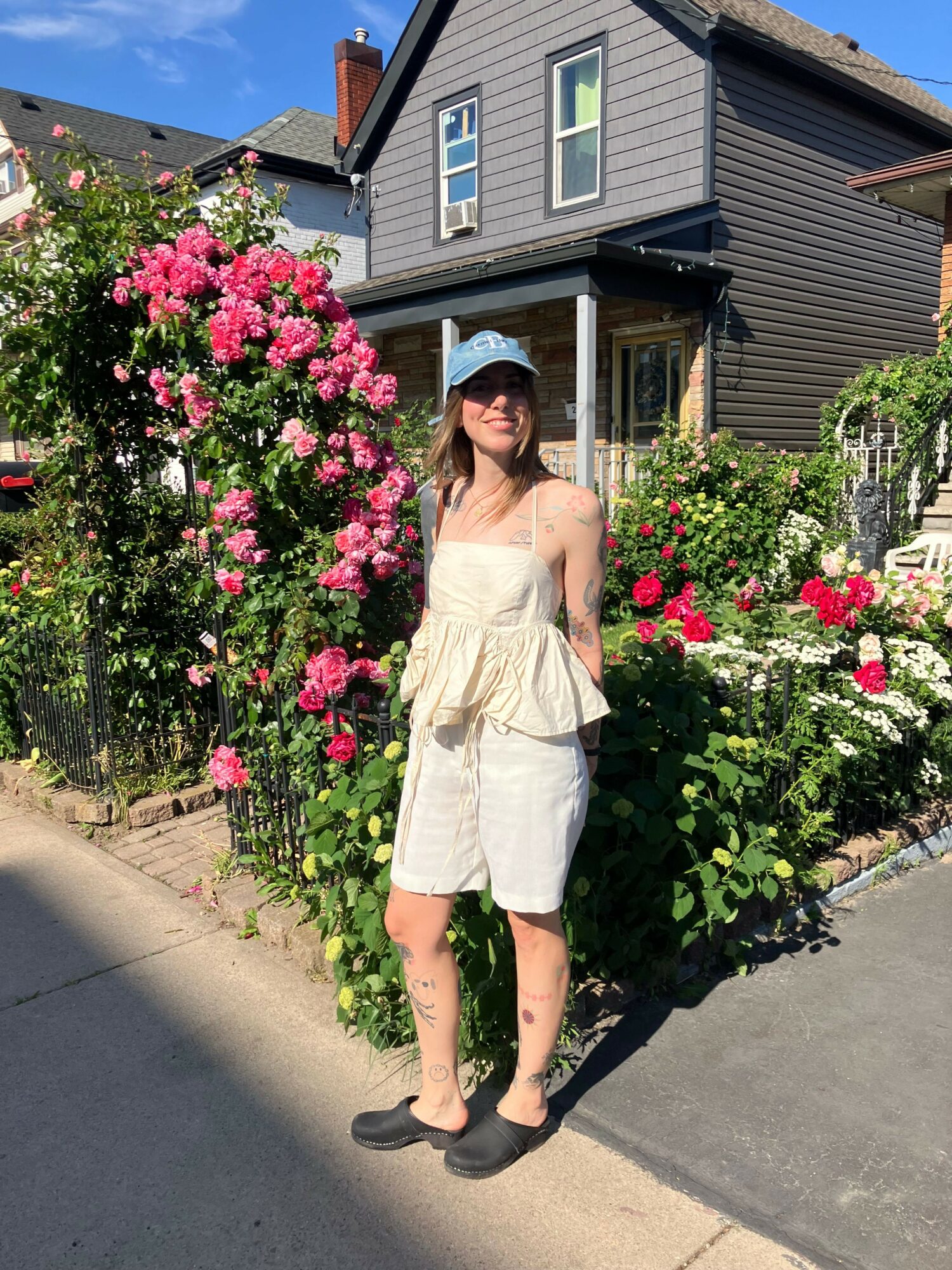 Person standing next to pink and red flowering bushes outside a house, wearing a beige dress and black shoes.