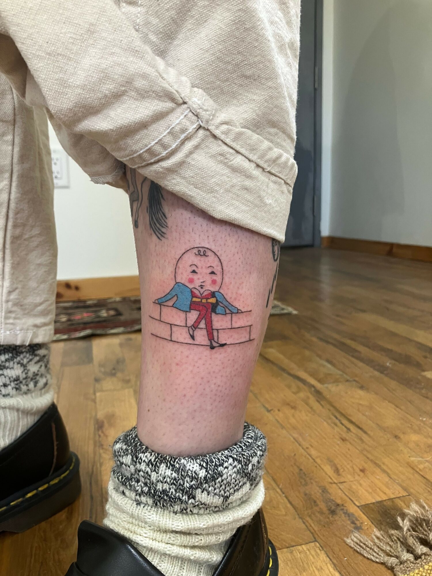 Tattoo of a person with a pink face, blue shirt, and red hair, sitting on a bench, on a person's leg.