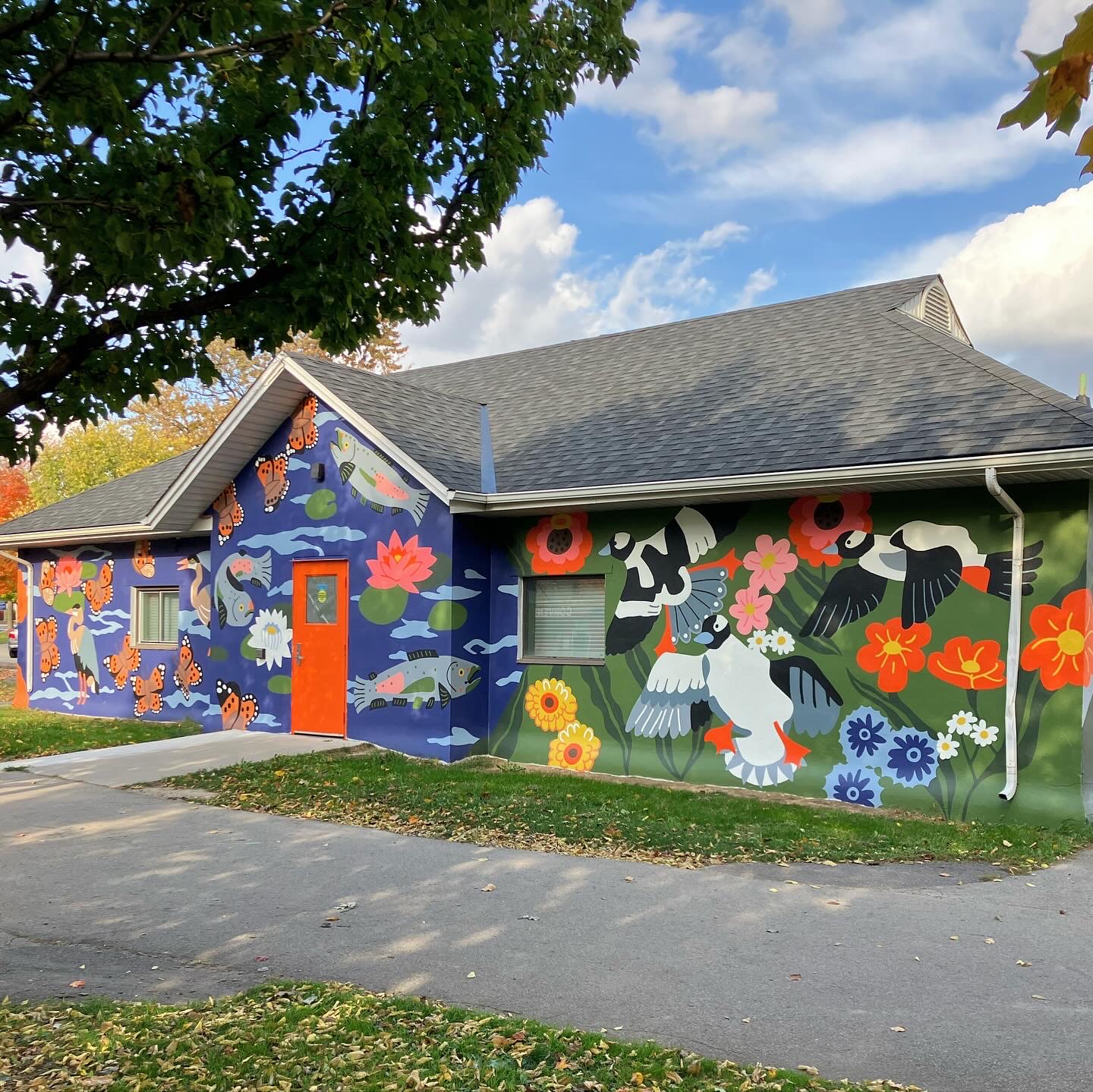 Building with colorful mural featuring flowers, animals, and abstract designs, under a partly cloudy sky, with a tree in foreground.
