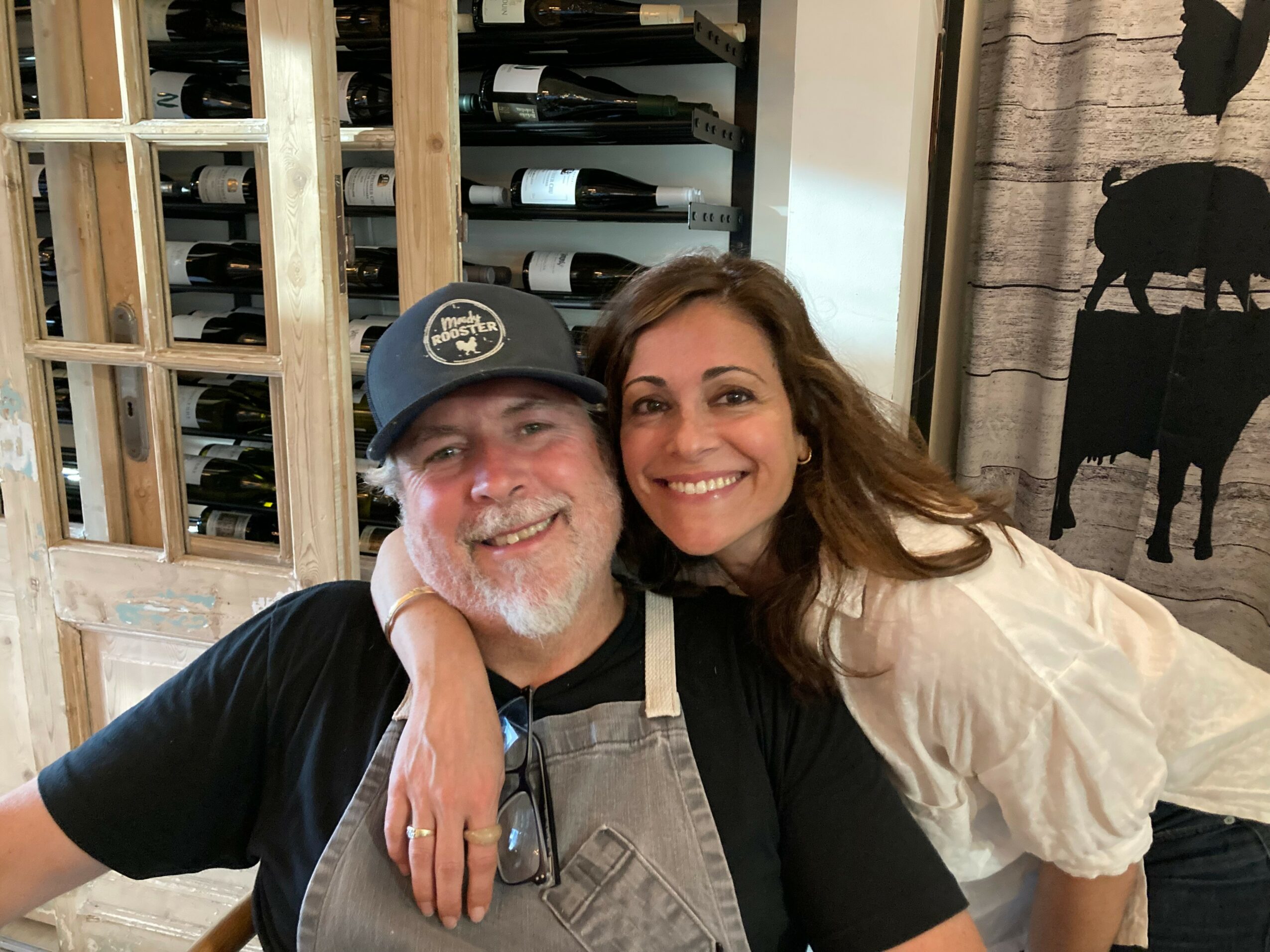 A smiling woman with long brown hair and a man with a beard and cap pose together indoors, with wine bottles in the background.