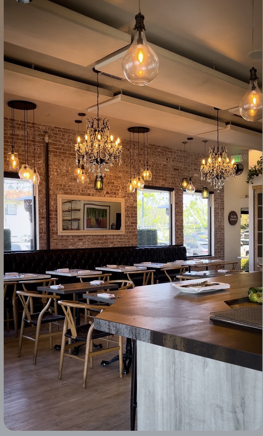 Interior of a restaurant with chandeliers, pendant lights, brick wall, tables, and chairs, illuminated by natural and artificial light.