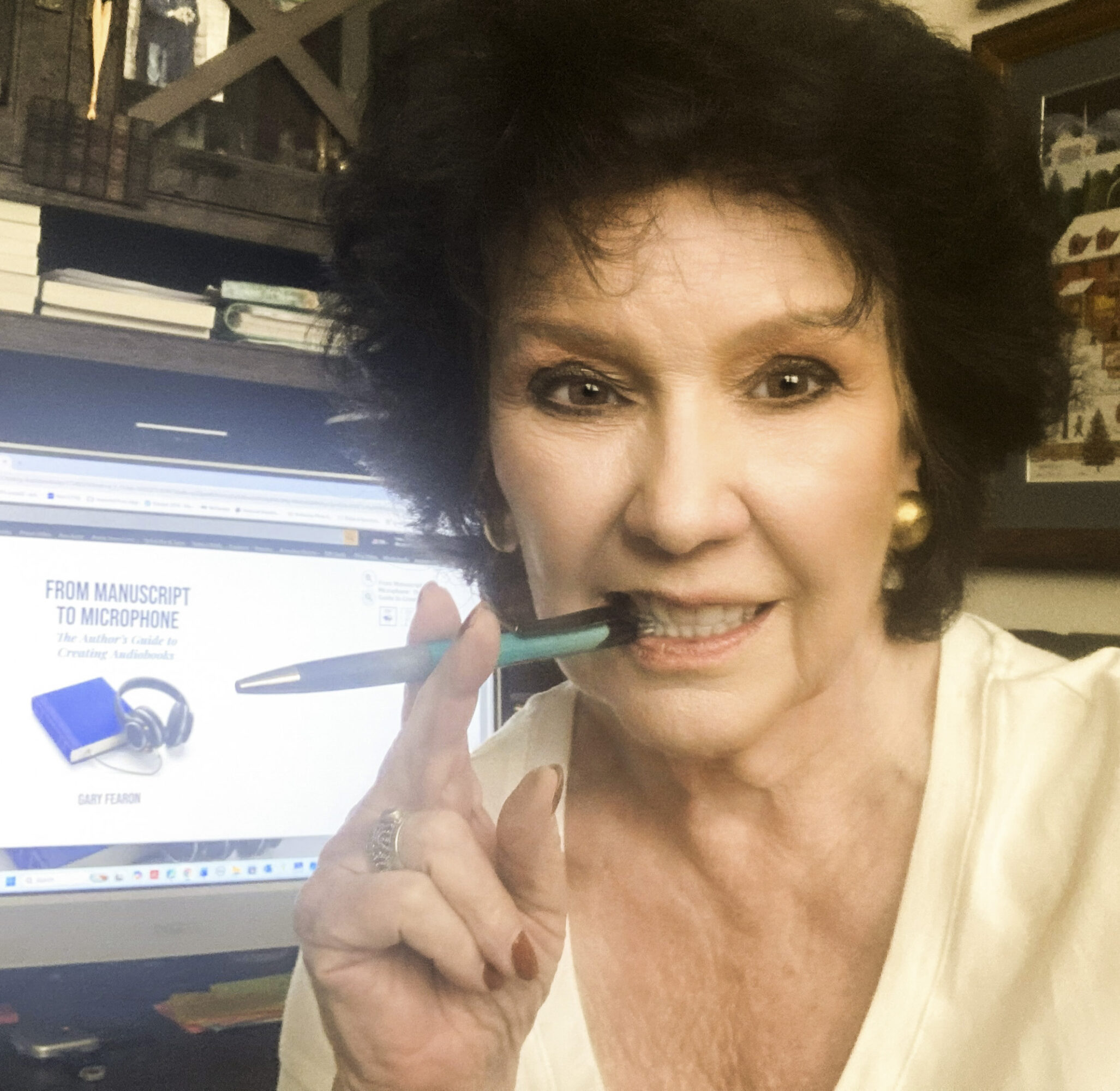 Woman with dark curly hair holding a pen near her mouth, smiling, in front of a computer screen with headphones image.
