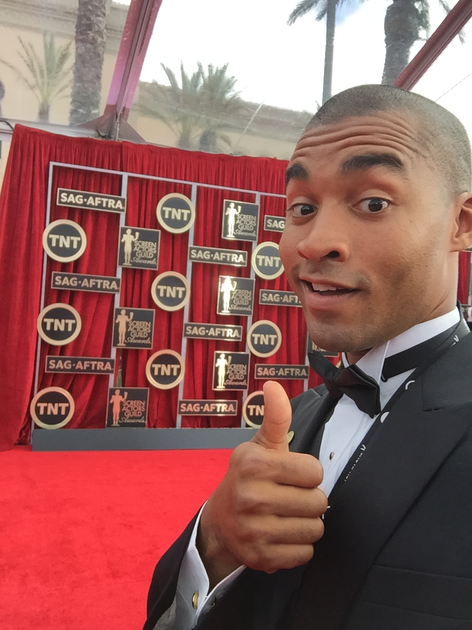 Man in tuxedo giving a thumbs-up in front of a red display with logos and images.