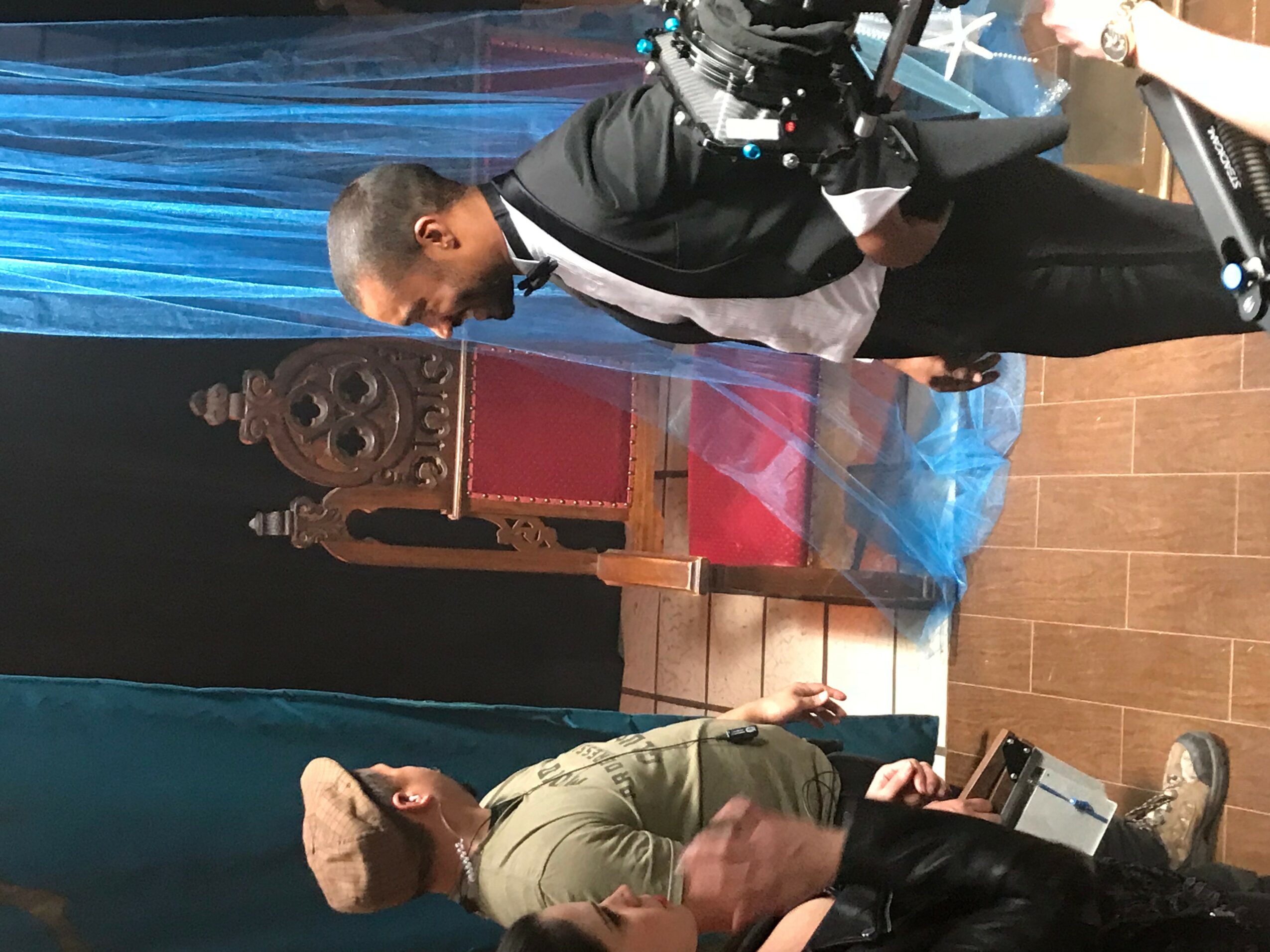 Two people, one in a suit and one in casual clothing, are in a room with a decorated backdrop and a wooden floor.