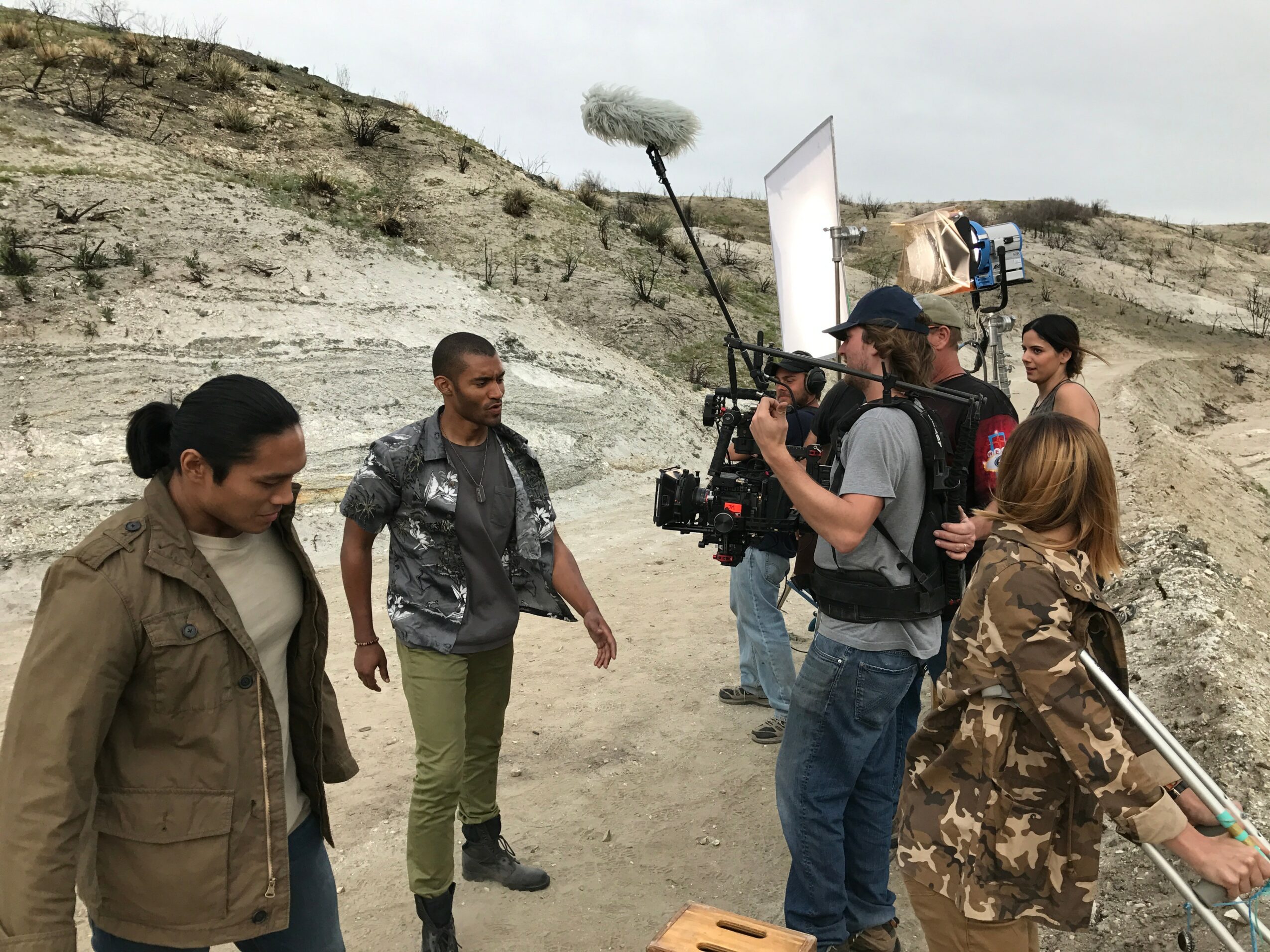Filming crew with camera and equipment filming two women outdoors on a dirt path with hills in background.