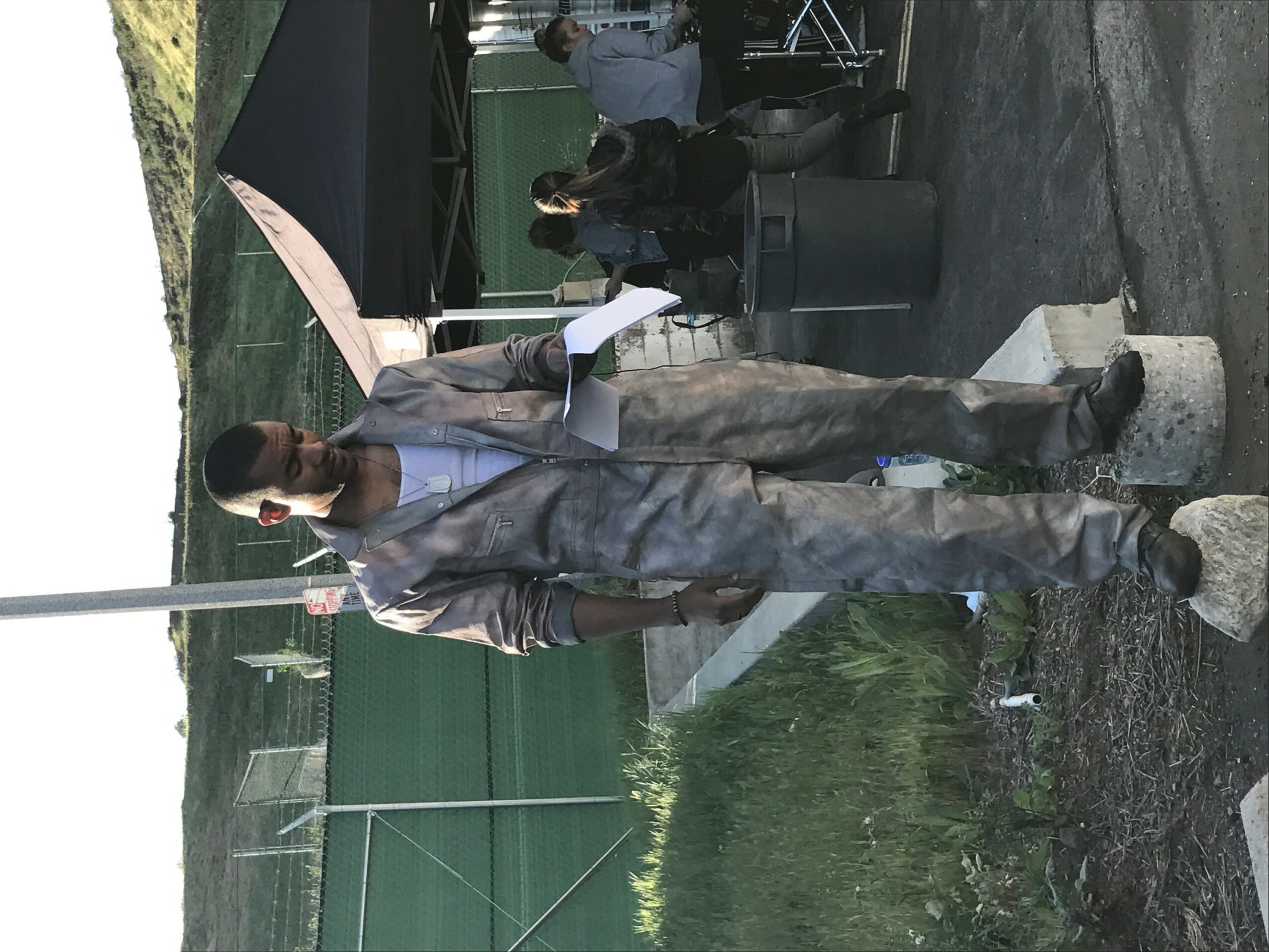 Person standing outdoors near a body of water, wearing a suit and holding a phone, with people and equipment in the background.