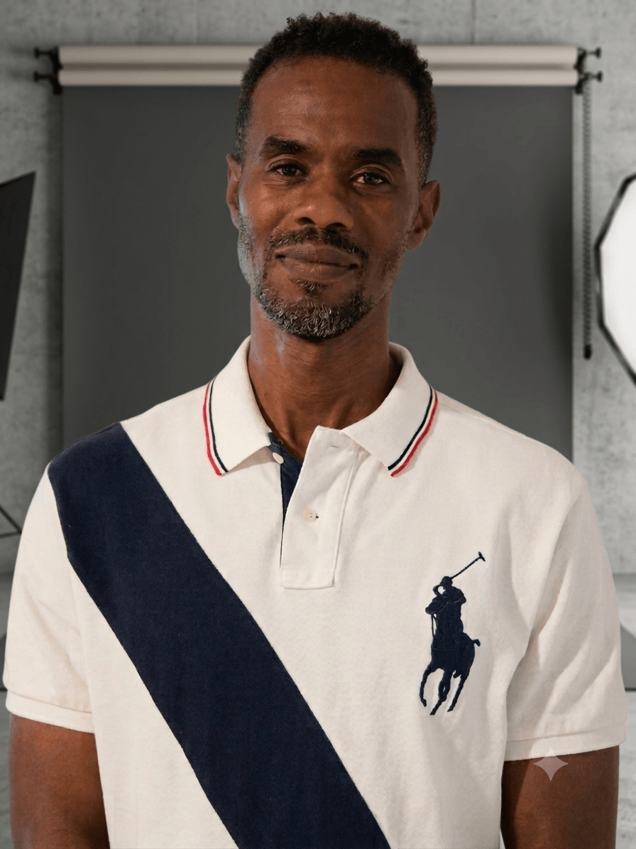 Man with short hair and beard wearing a white polo shirt with navy blue diagonal stripe and logo, standing indoors.