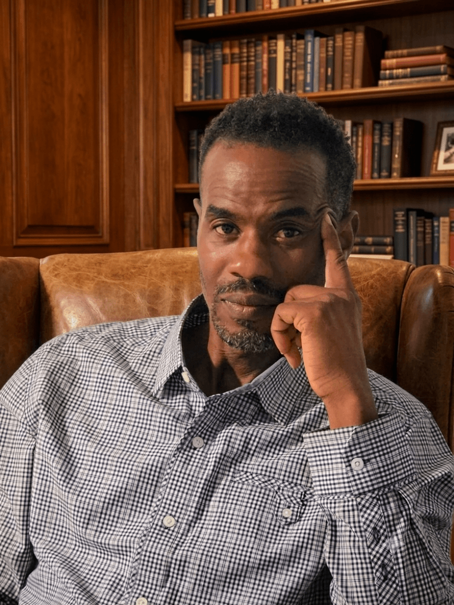 Man with short curly hair and a beard, wearing a checkered shirt, sitting in a room with wooden bookshelves filled with books, resting his head on his hand.
