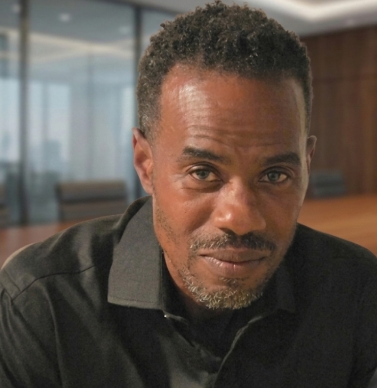A man with short curly hair and a beard, wearing a black shirt, sitting in a modern office setting.