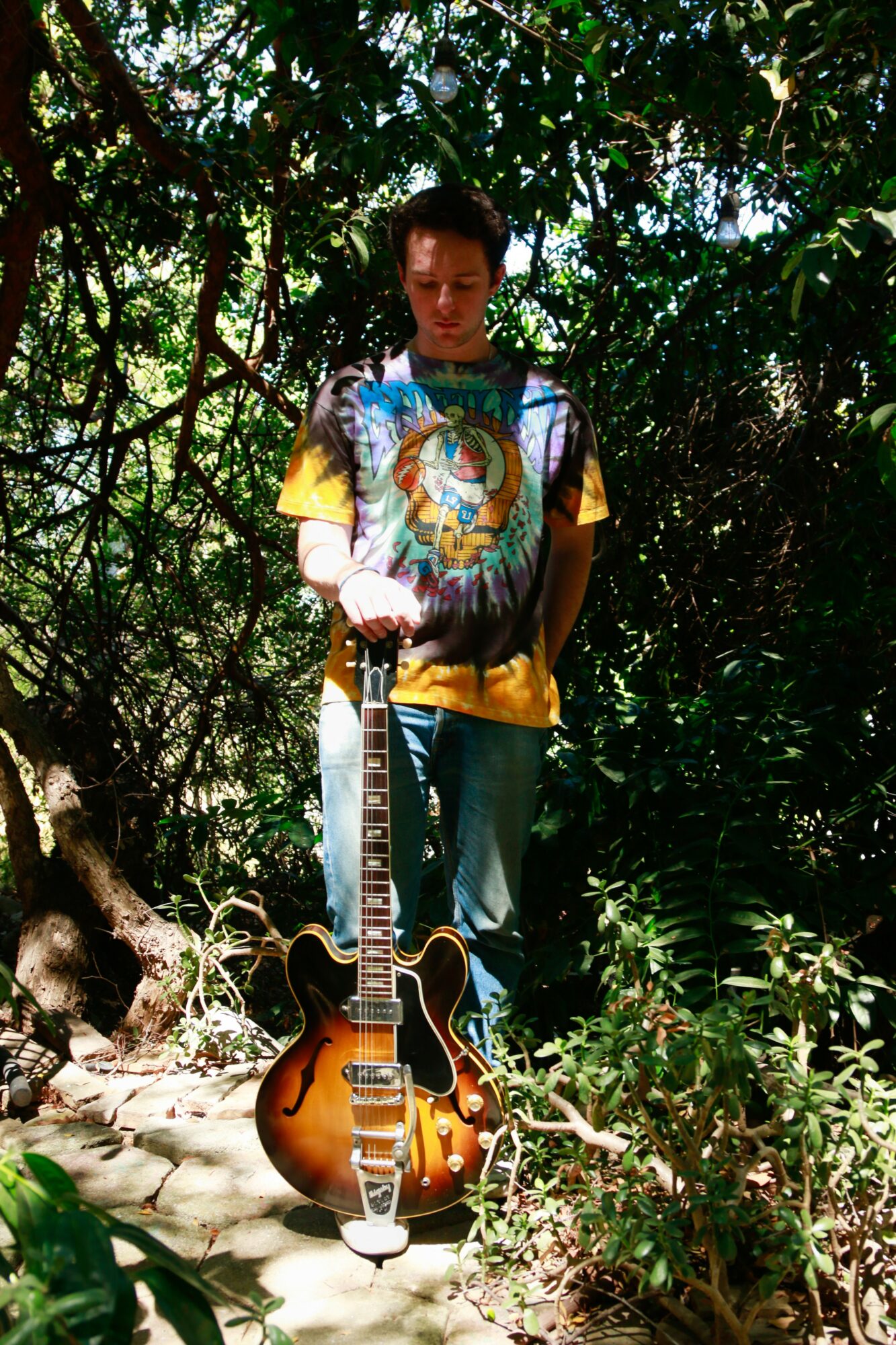 Young man standing outdoors among trees with an acoustic guitar on the ground in front of him.