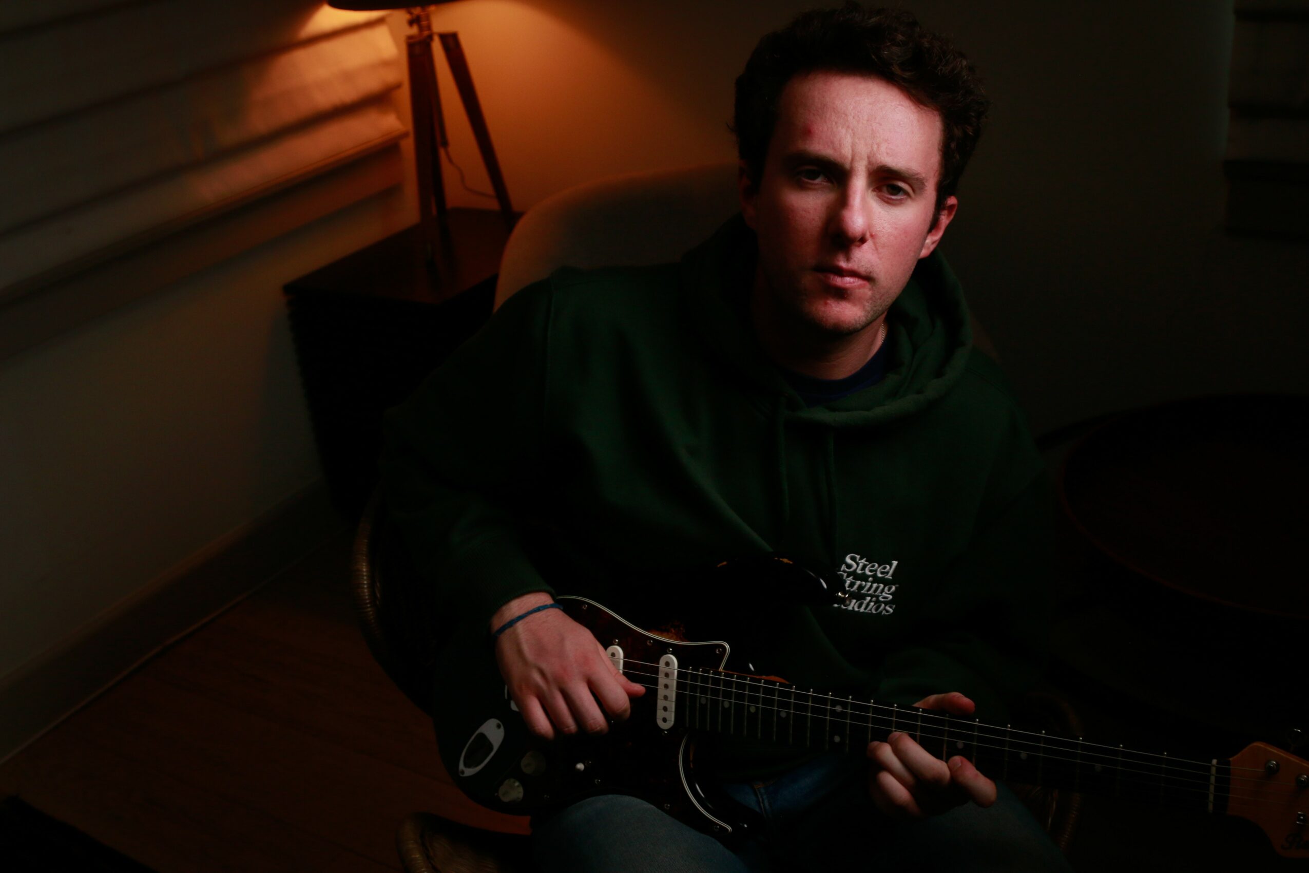 Person sitting in a dimly lit room holding a guitar, with a lamp in the background.