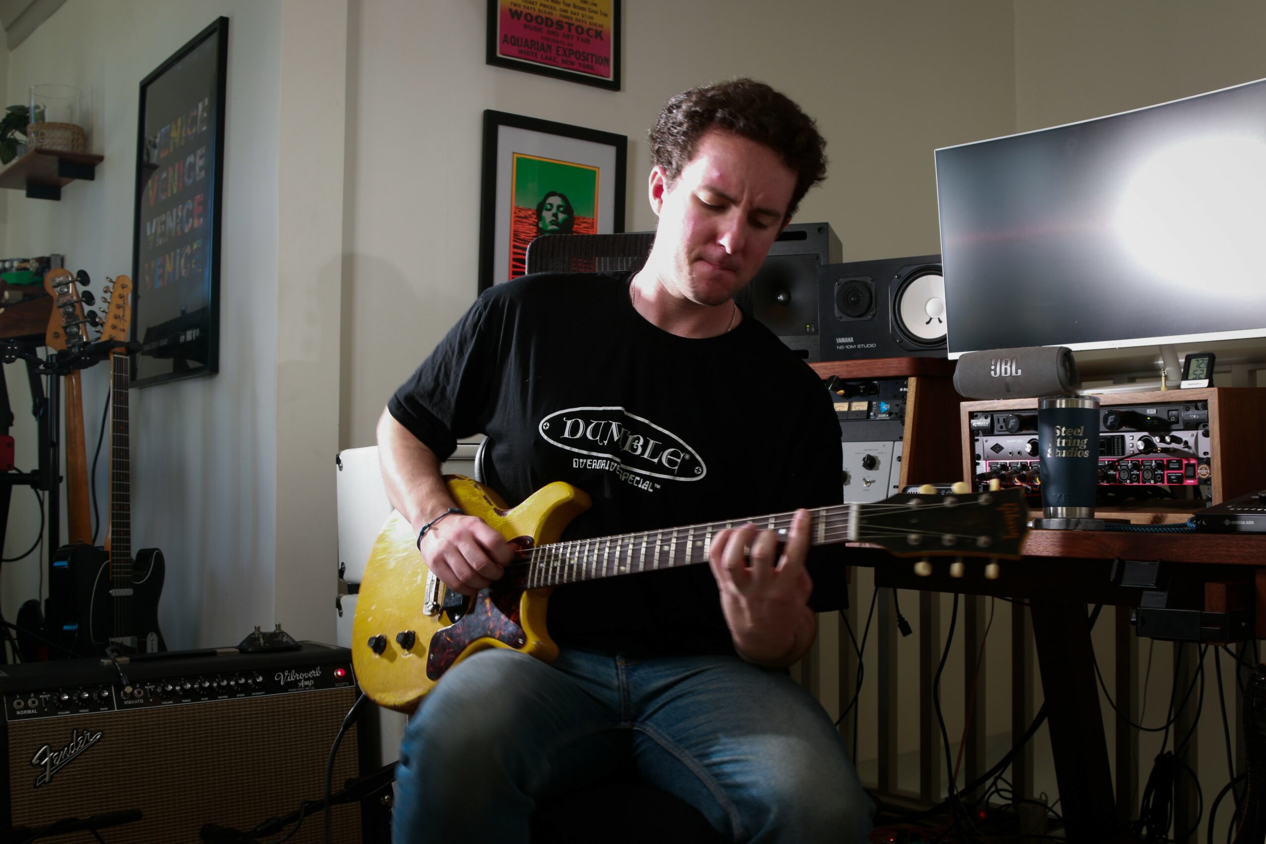Person playing yellow electric guitar in a music studio with equipment and framed pictures on the wall.