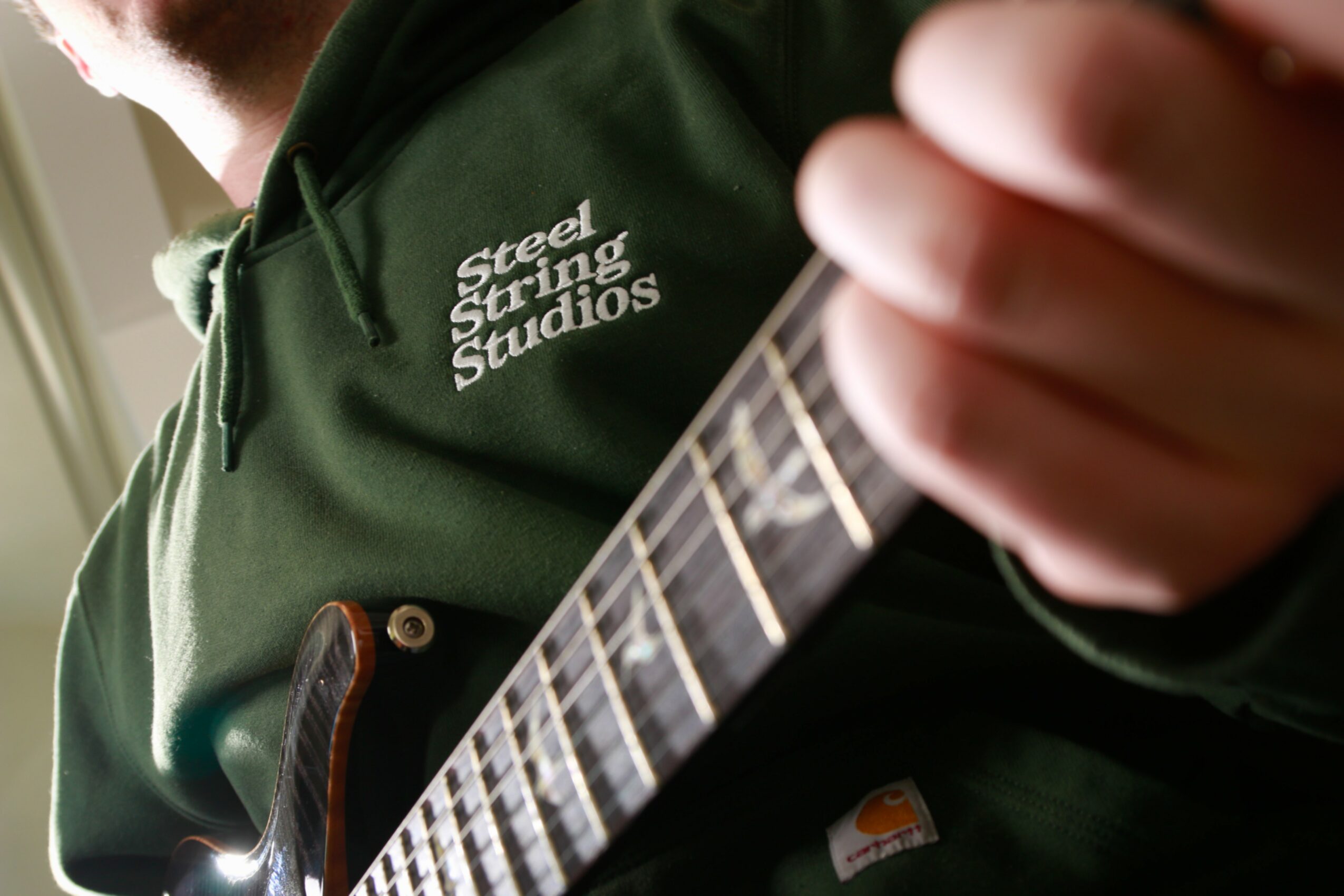 Person wearing a green hoodie playing an electric guitar, focusing on the hand and guitar neck.