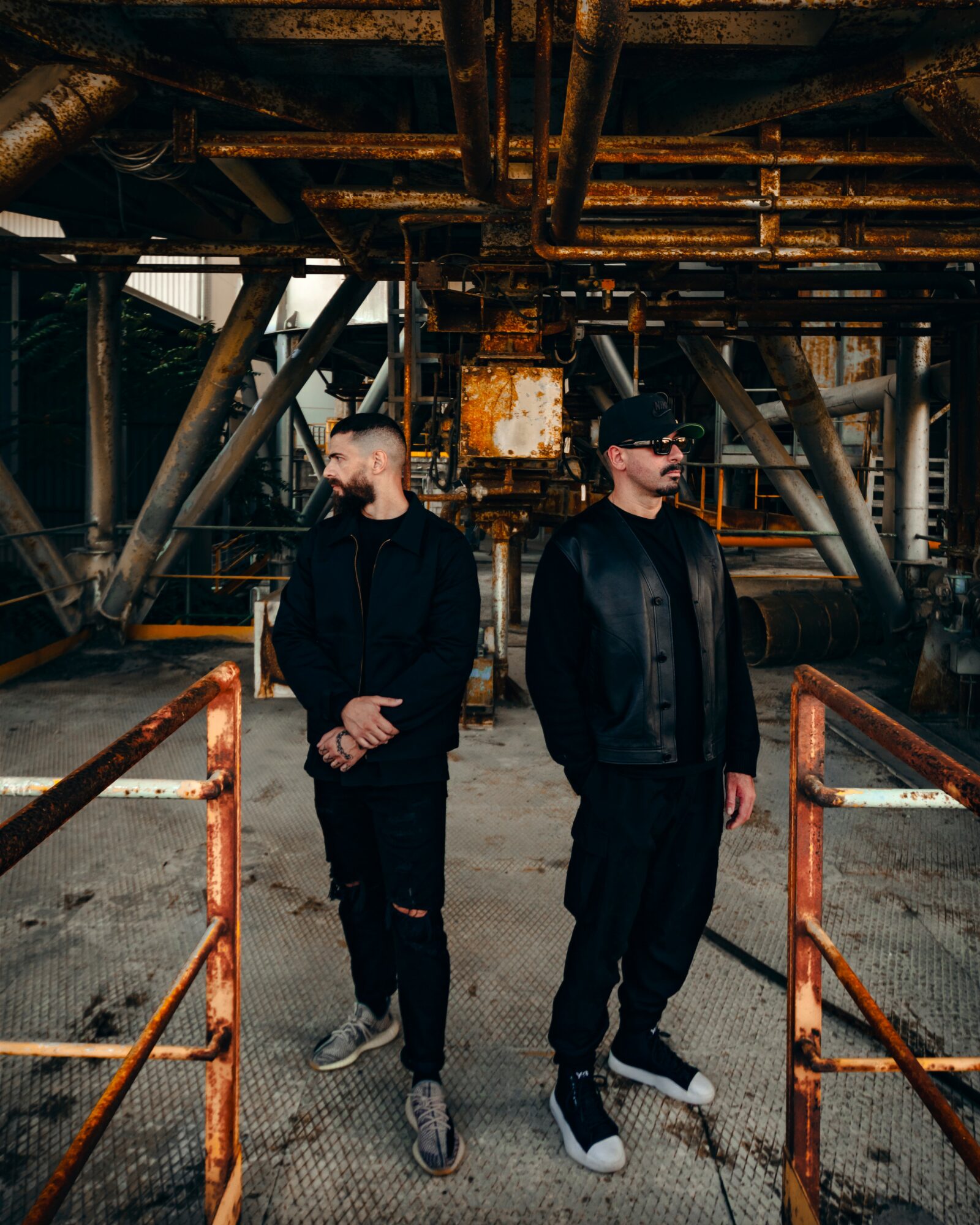 Two people stand in an industrial setting with metal pipes and structures, one with hands clasped, the other with hands in pockets.