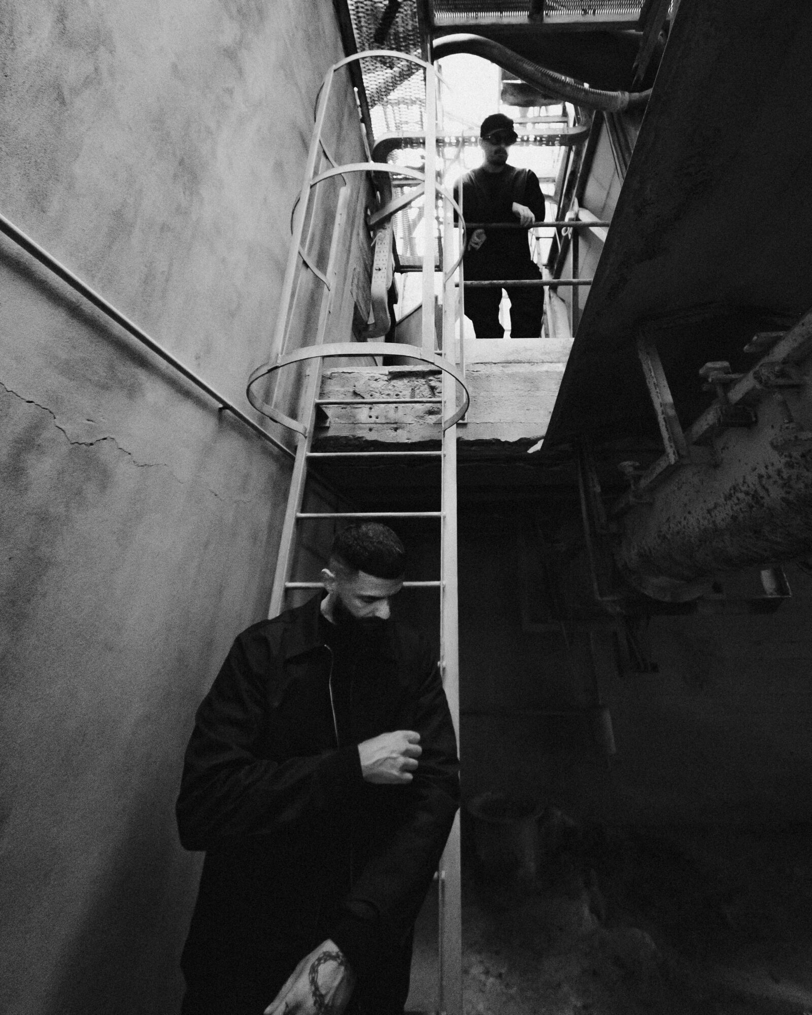 Person in dark clothing looking down, ladder and another person visible in background, black and white photo.