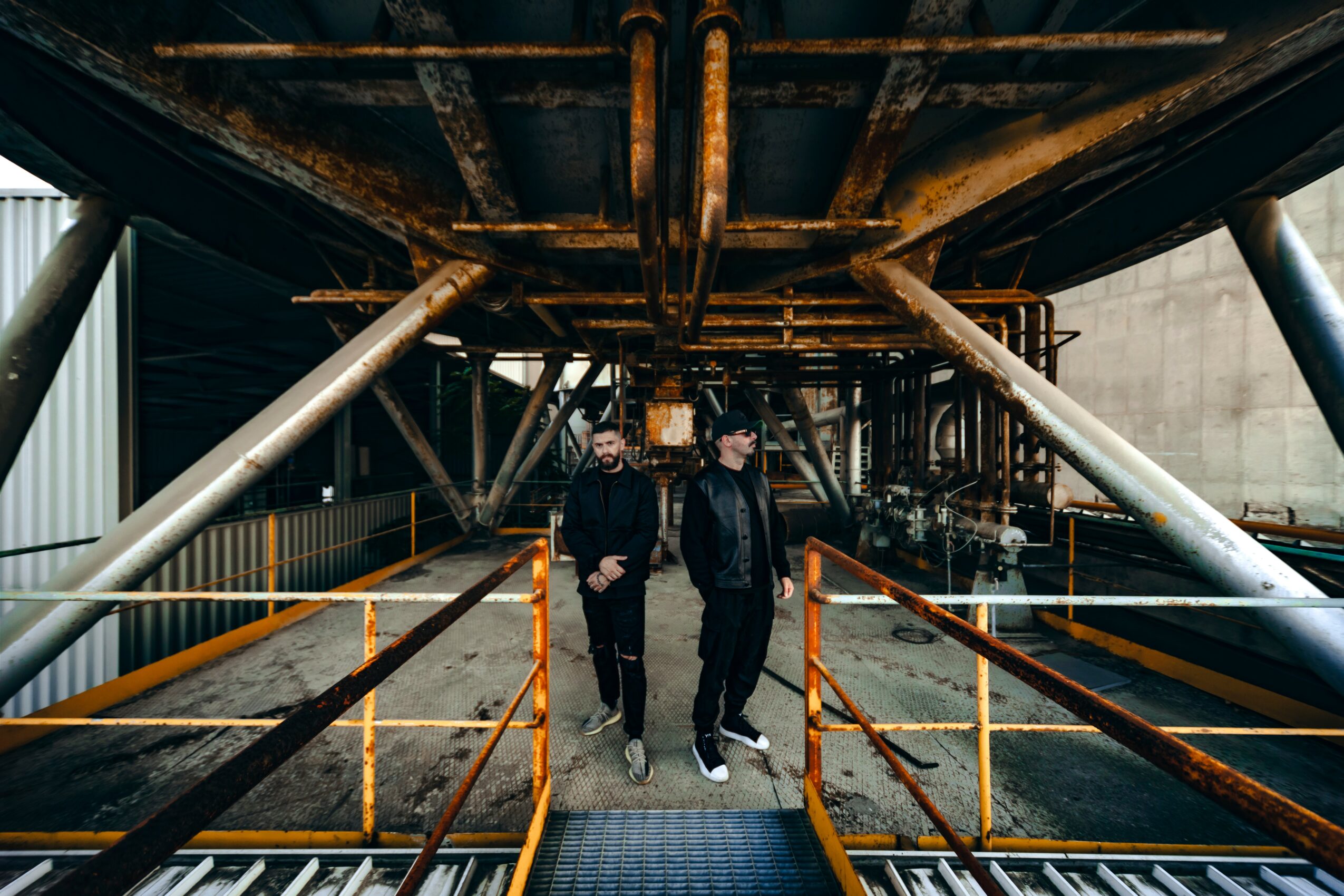 Two people walking under a large industrial structure with metal beams and orange railings.