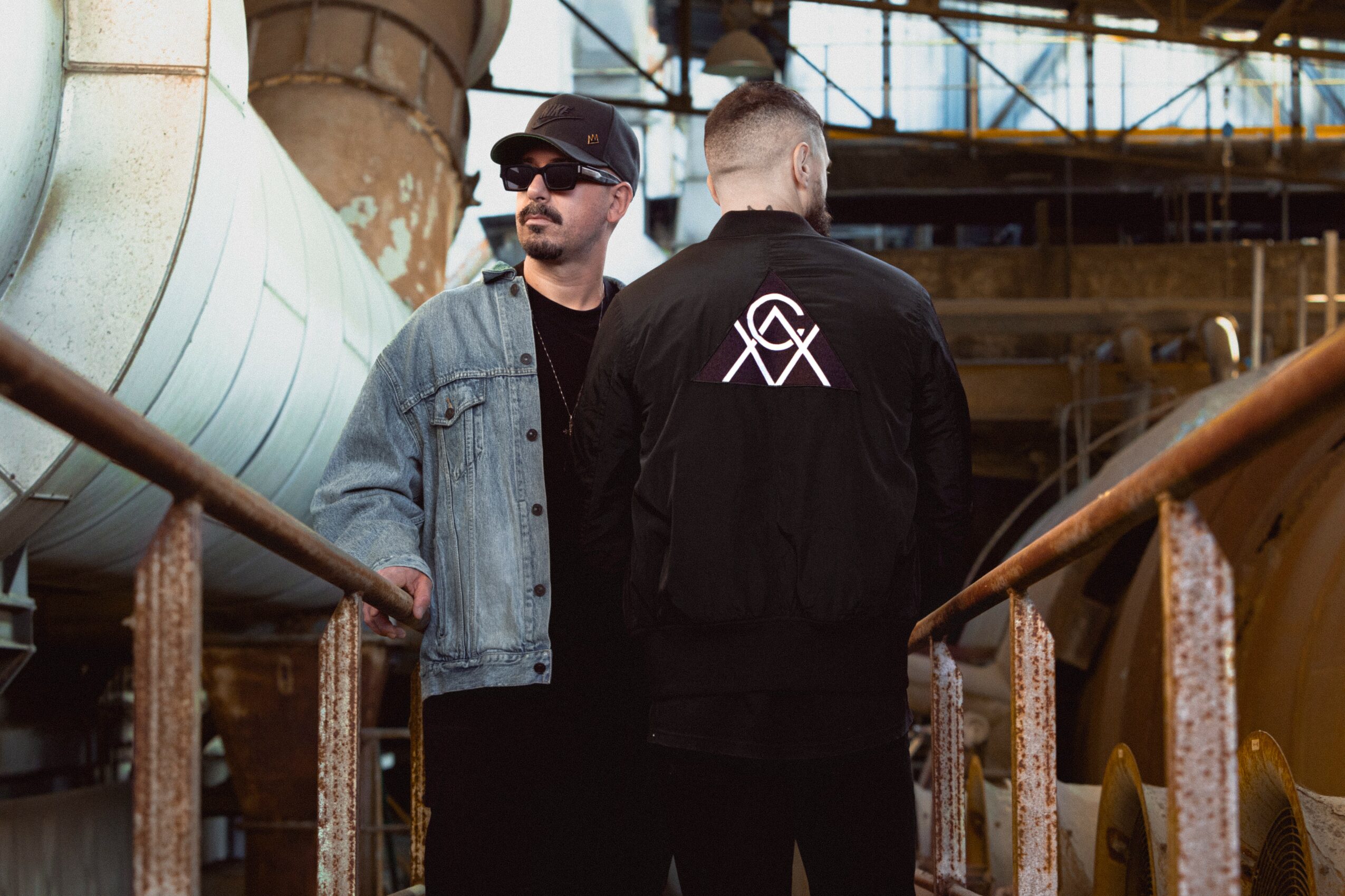 Two men stand on an industrial ship deck with pipes and metal structures in the background.