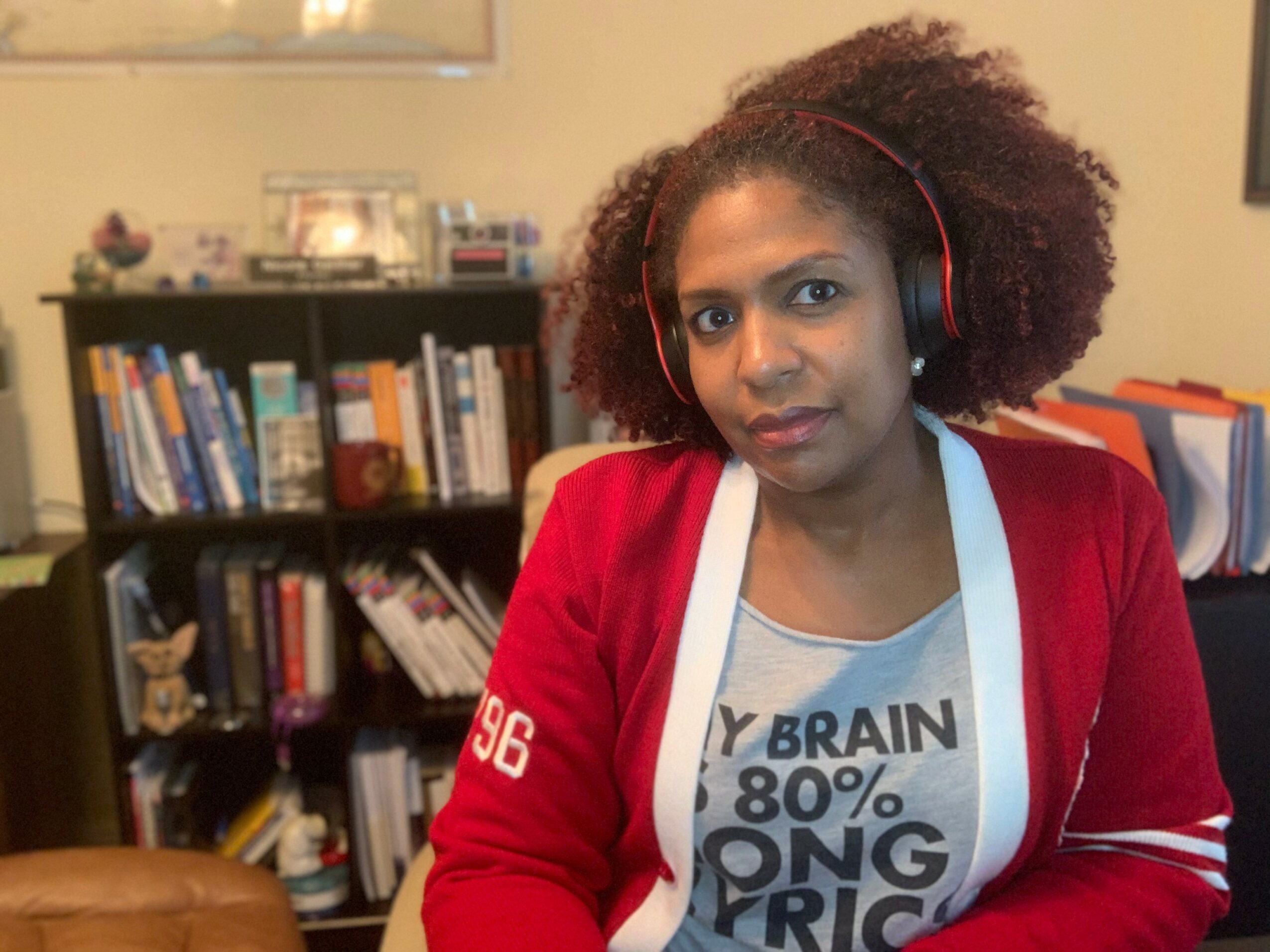 Woman with curly hair wearing headphones, red jacket, and gray T-shirt in a room with bookshelves.