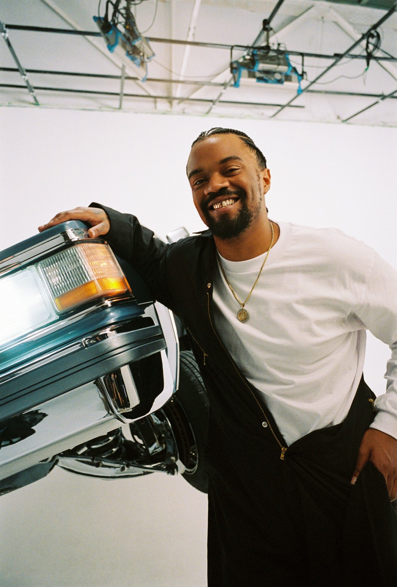 Smiling man with a beard and gold chain leaning on a car in a studio with a white background.