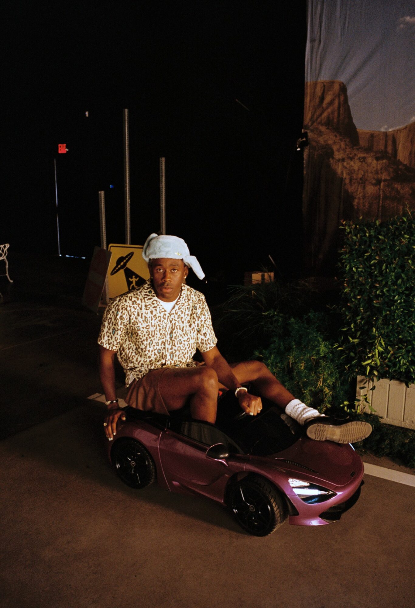 Child sitting in a small toy car, wearing a white cap and patterned shirt, in a dark outdoor setting with plants and a sign.