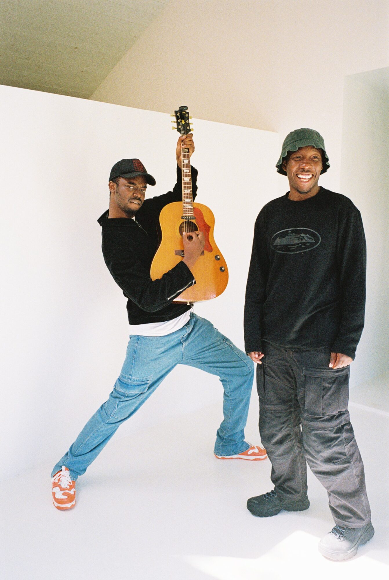 Two men stand against a white background, one holding a guitar and the other smiling, wearing casual clothing.