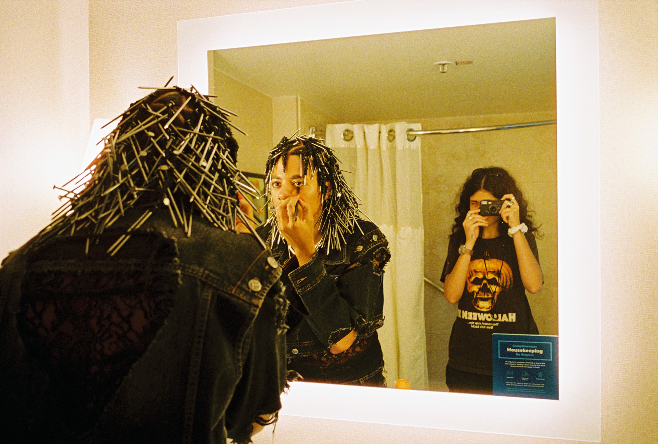 Two people with dreadlocks looking at a mirror, one covering their face, another taking a photo in a bathroom.