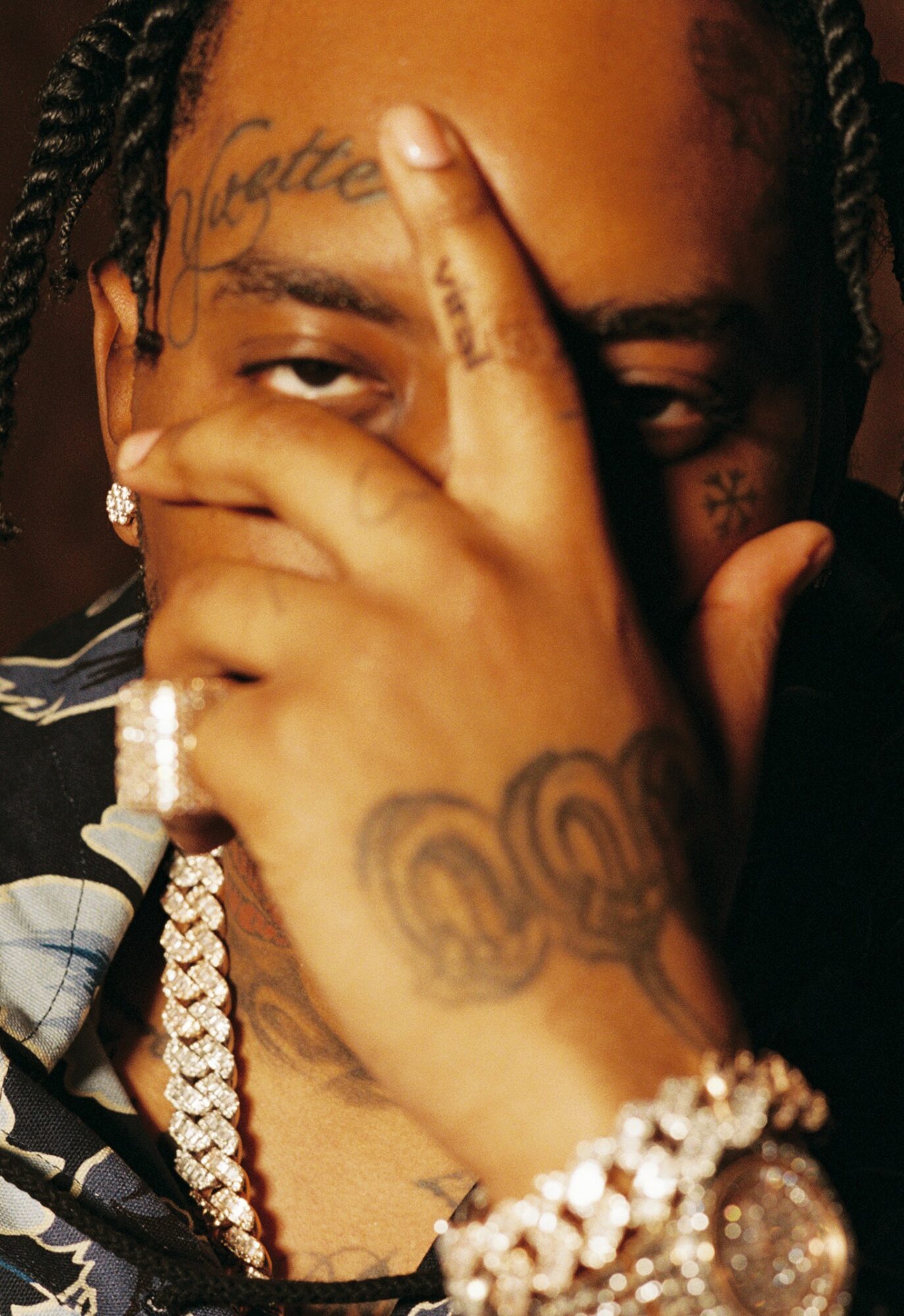 Person with tattoos covering face and hand, wearing jewelry, holding hand near face, with dark background.