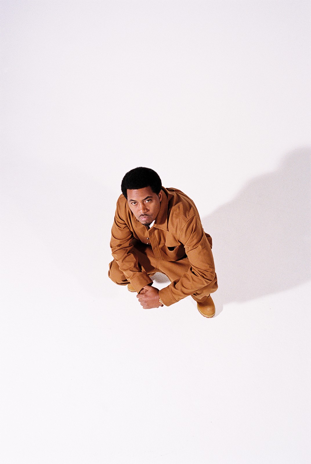 Person crouching on white background, looking up, wearing brown outfit, shadow cast to the right.