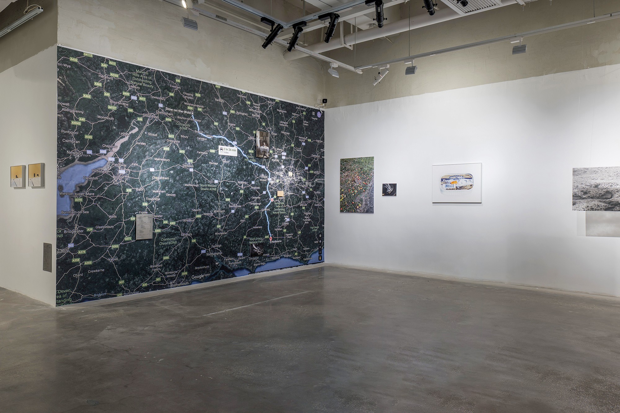 Gallery with a large map on the left wall and several smaller artworks on the right wall, empty floor, white walls.