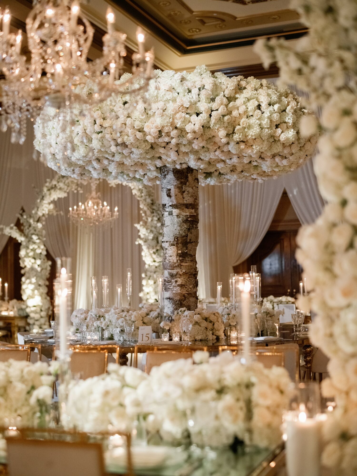 Elegant event with large floral centerpiece on a tall stand, surrounded by candles and floral arrangements, in a decorated hall.
