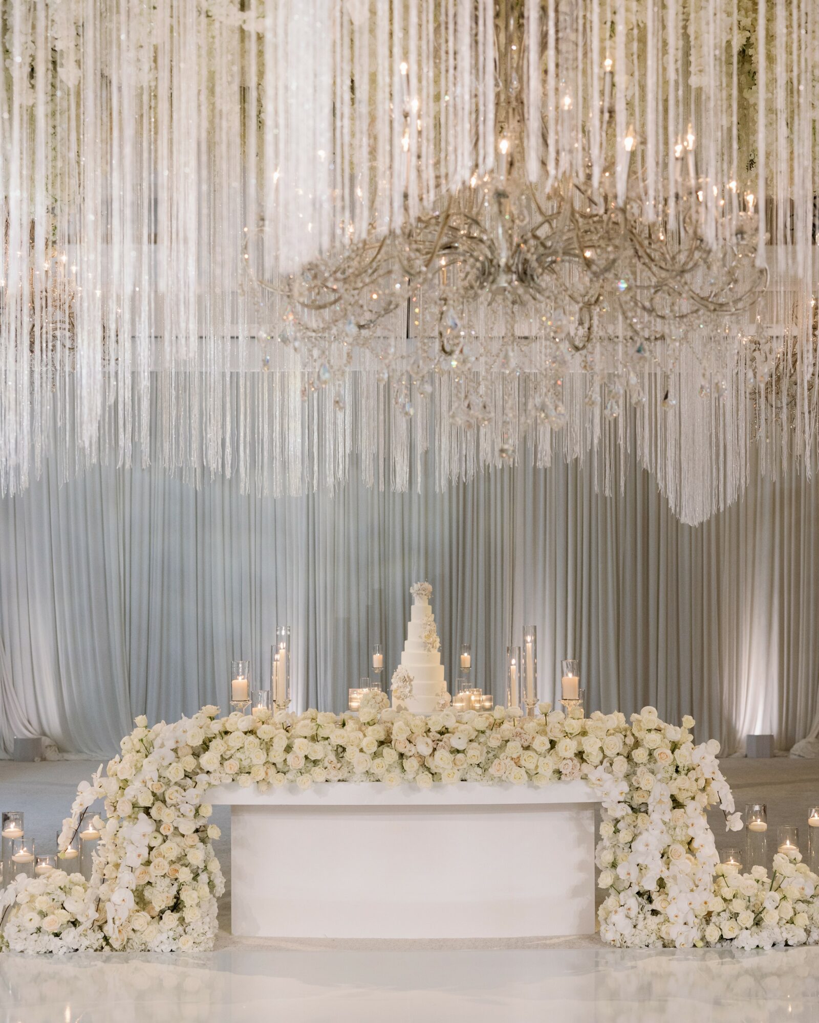 Elegant event setup with a floral arrangement, candles, and a chandelier, against a draped backdrop.