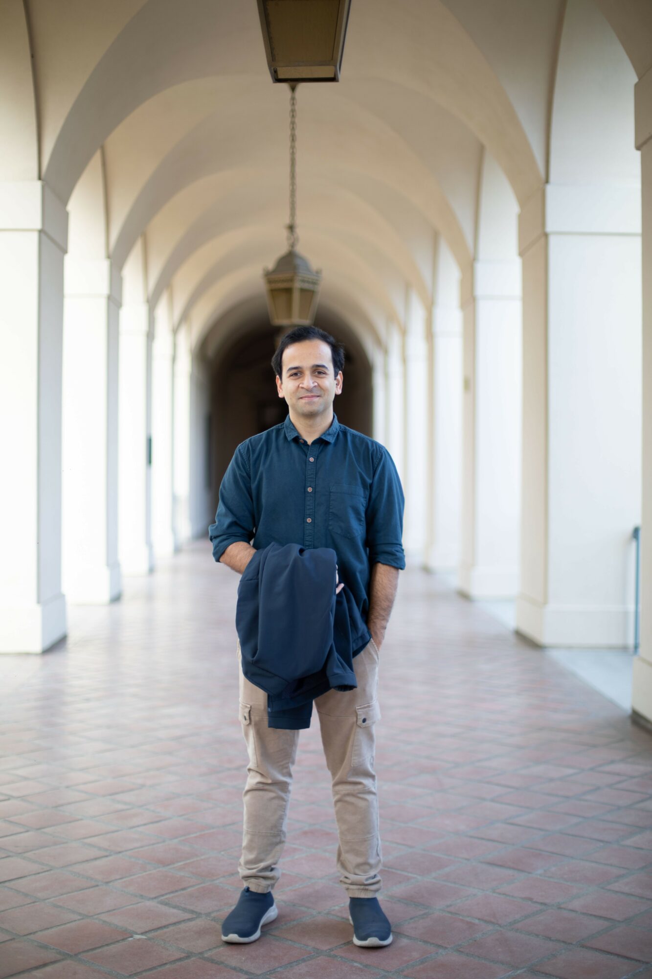 Young man standing in a corridor with arches, holding a jacket, smiling, natural light, tiled floor.