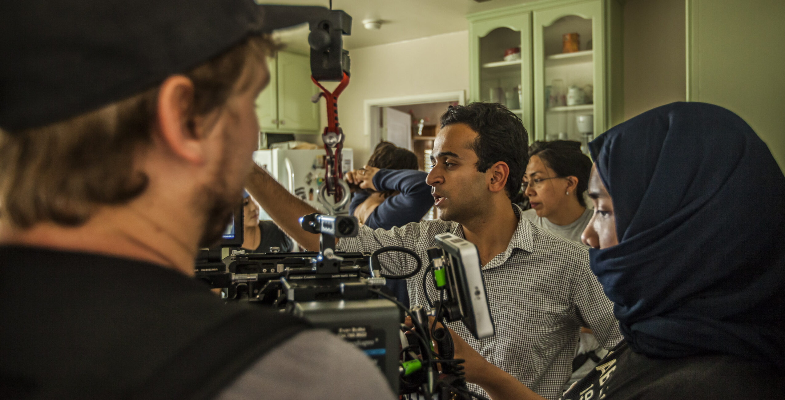Group of people working with camera equipment in a room with green cabinets, some wearing glasses and head coverings.