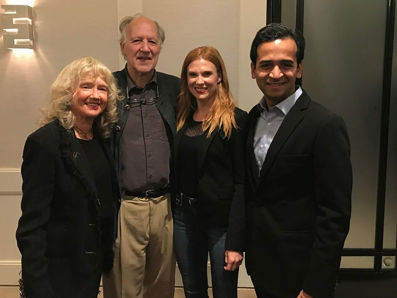 Four people standing together indoors, smiling, with a beige wall and elevator behind them.