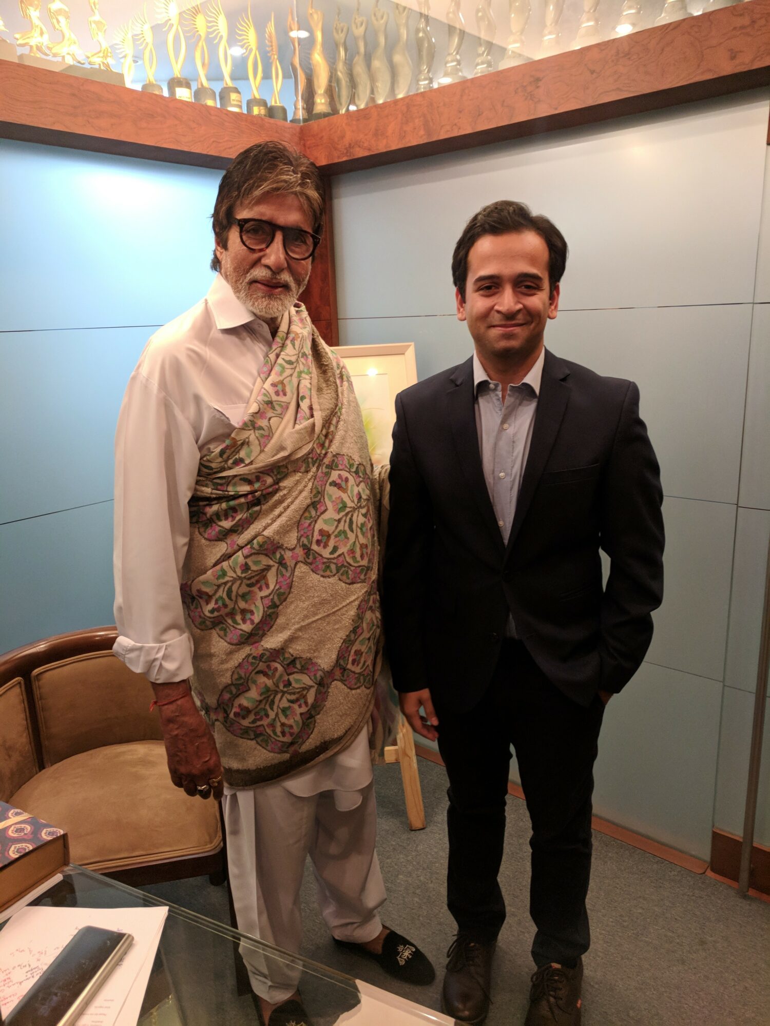 Two men standing indoors, one older with glasses and a patterned vest, the other younger in a suit, smiling.