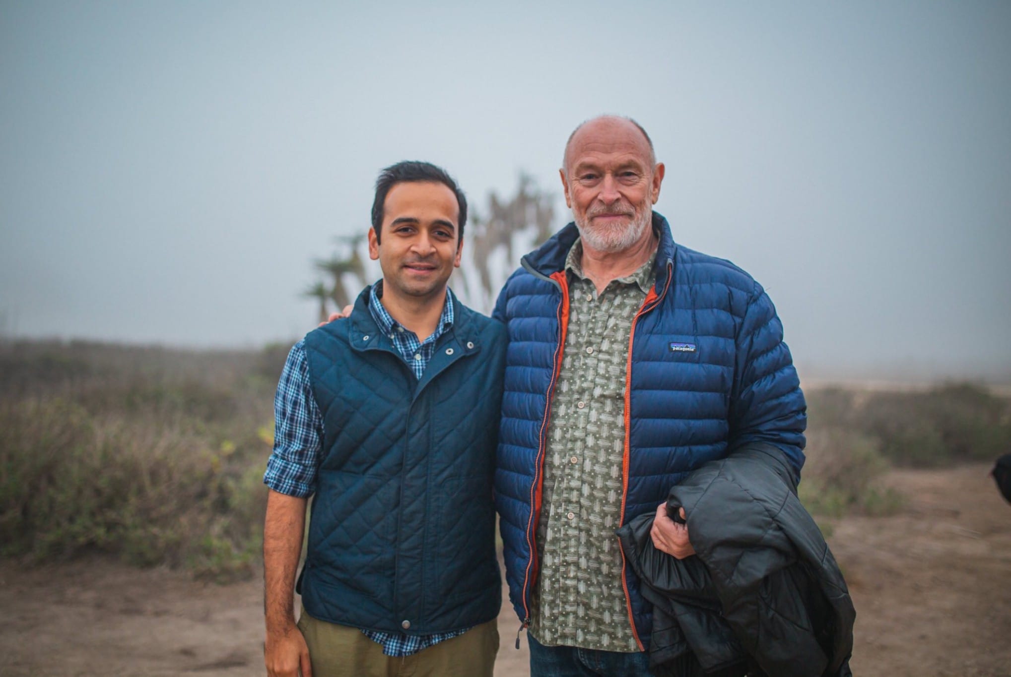 Two men standing outdoors on a cloudy day, one younger and one older, both wearing jackets, with a landscape background.