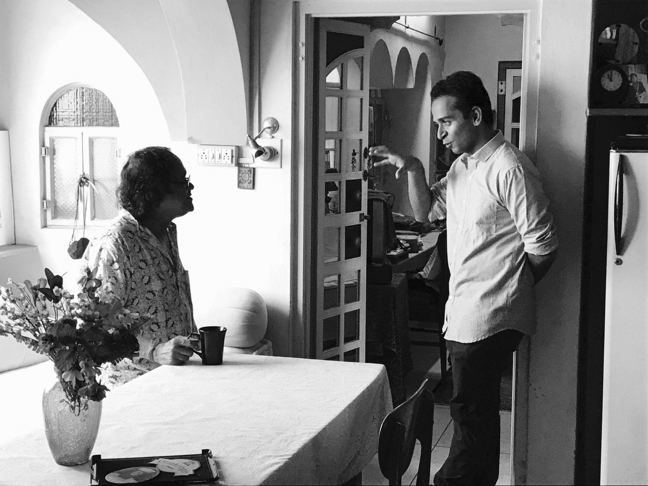 A young man and an elderly woman are in a kitchen, with the woman sitting at a table holding a mug, and the man standing near a doorway.