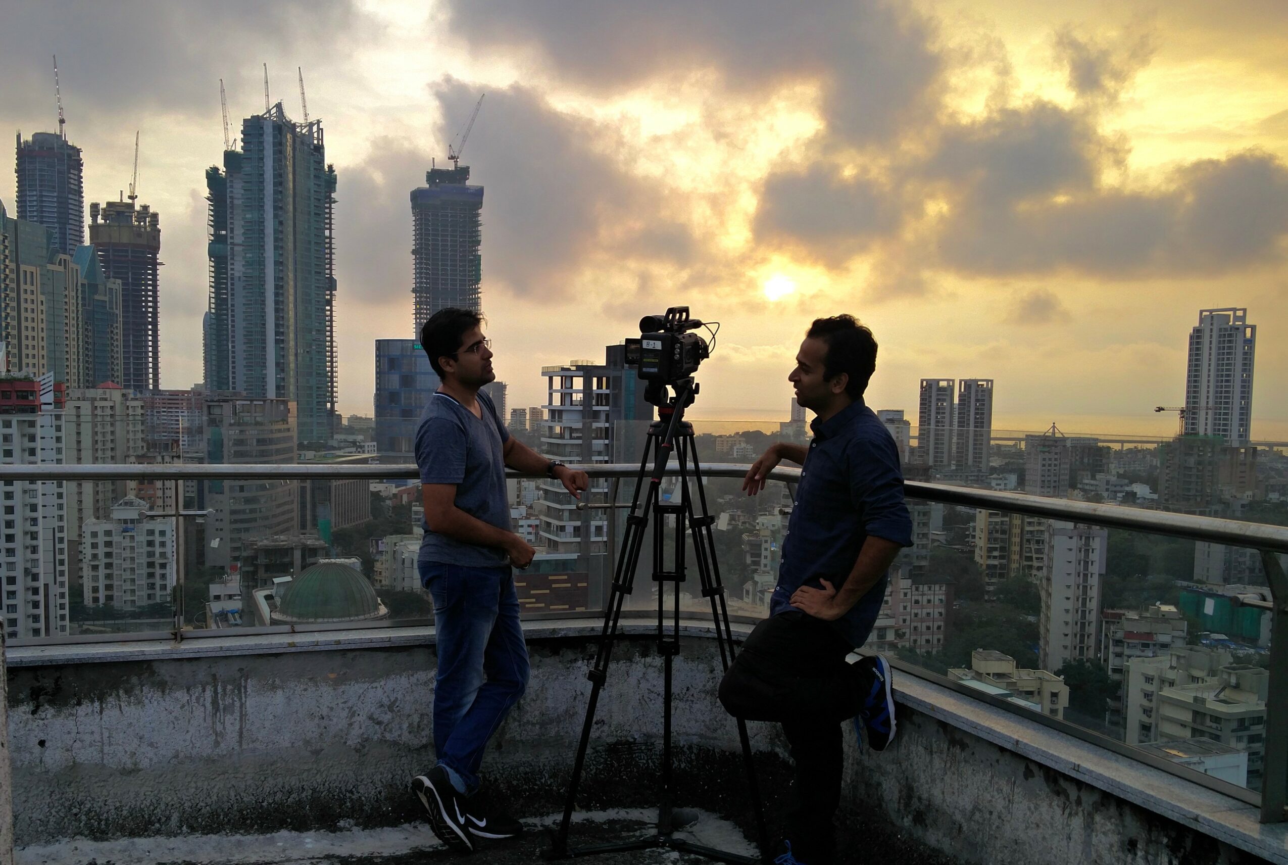 Two people filming on a rooftop with a city skyline and sunset in the background.