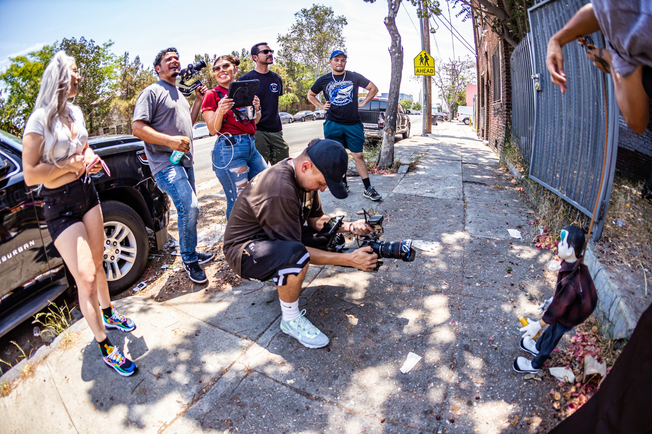 Group of people on sidewalk watching a young girl in a jacket and hat, with a camera operator nearby, outdoors.