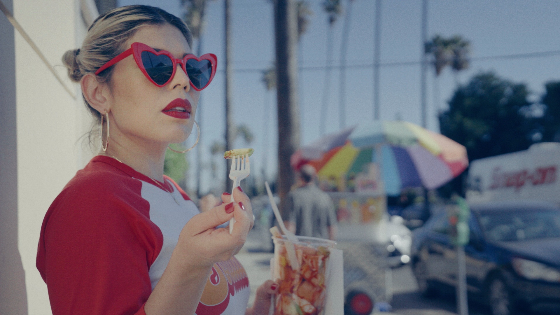 Woman with red heart-shaped sunglasses and red lipstick holding a drink and a fork outdoors.