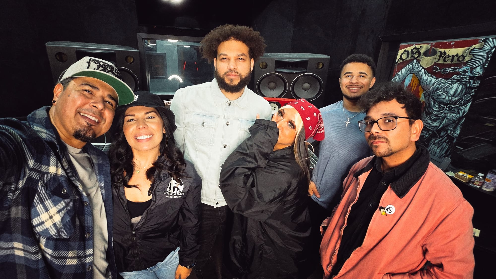 Group of six people smiling in a music studio with speakers and equipment behind them.